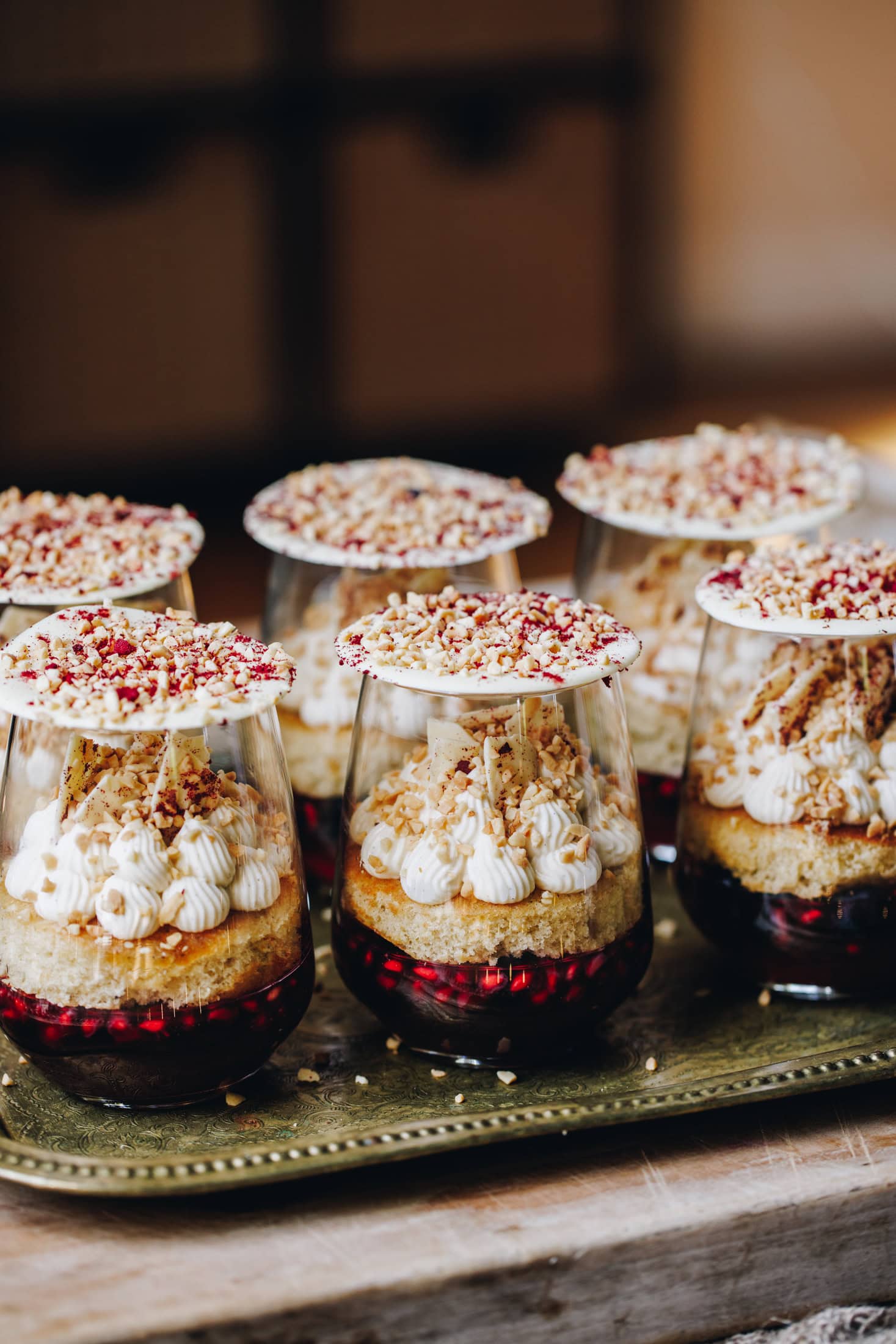 The mini berry trifles have had sponges added to them on top of the set jelly. Sponge has whipped cream piped on to it. The cream has white chocolate shards and chopped nuts on top and white chocolate discs sit over the glasses.