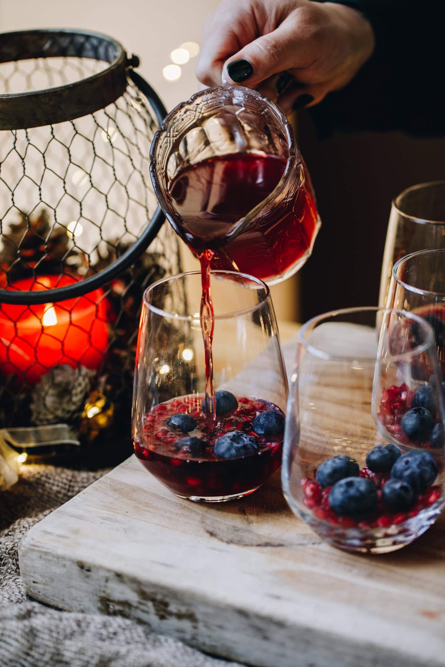 Glasses are on a wooden board and berries are inside glasses that are on the board. A glass vintage jug is pouring a cranberry jelly mixture in to the glasses. There is an orange candle in the background.