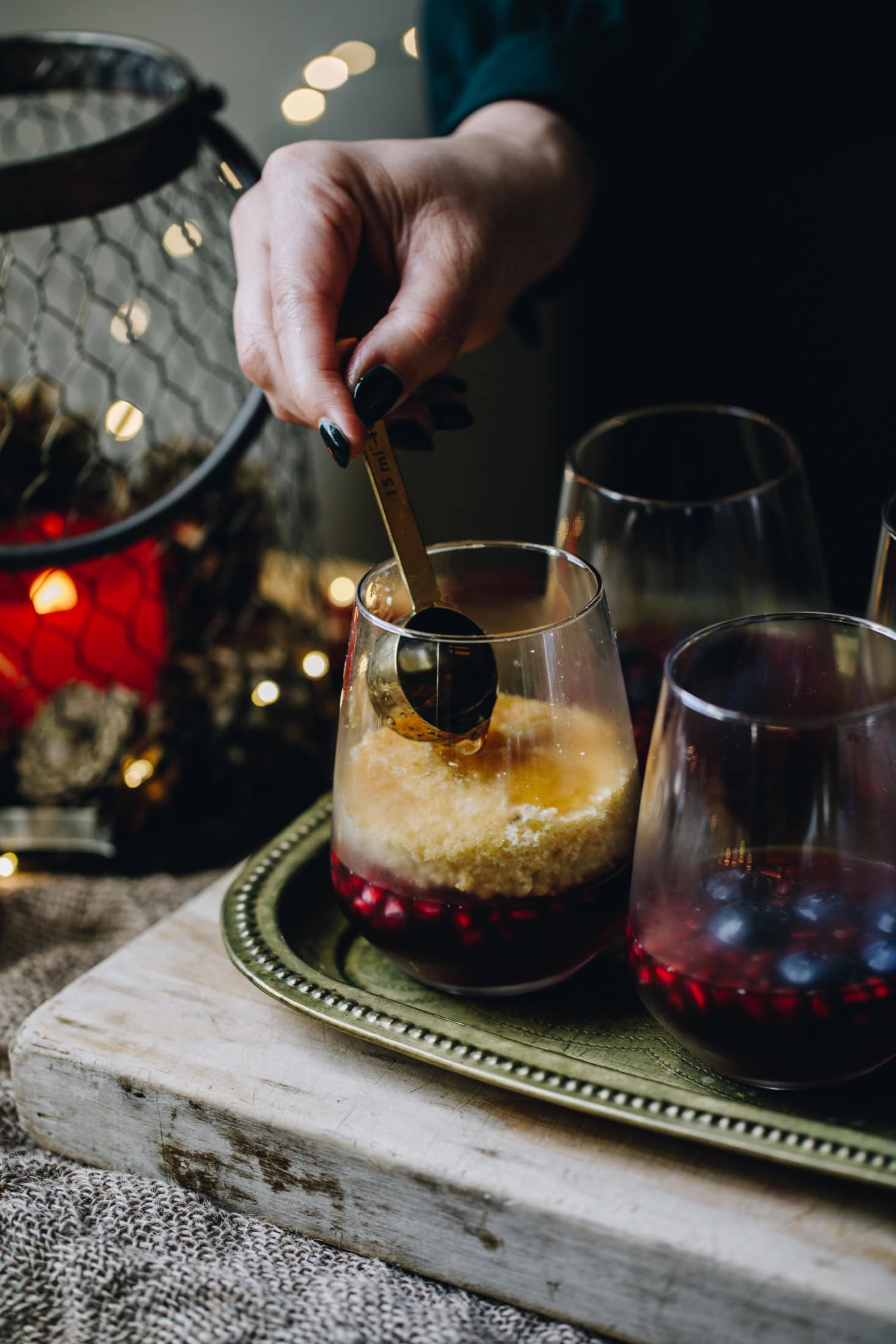 The mini berry trifles have had sponges added to them on top of the set jelly. A sponge is having a tablespoon of vanilla syrup added on to it. The glasses are on a gold tray.