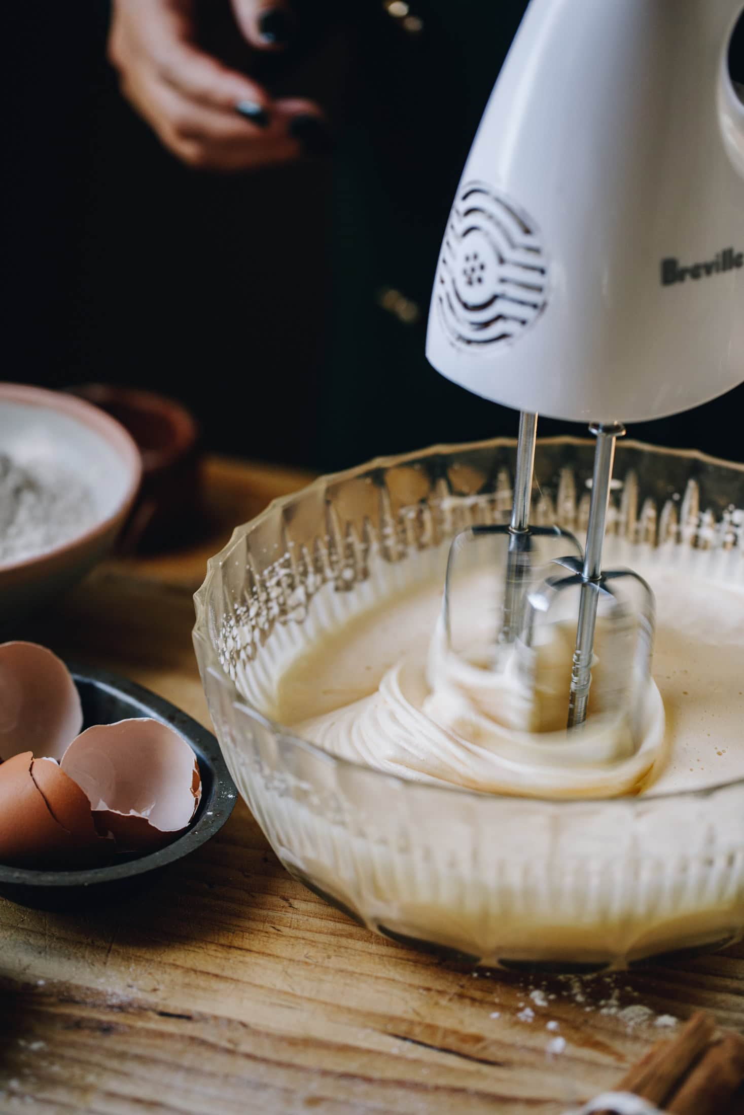 A vintage bowl is on a wooden table. Eggs are being whipped with a white hand mixer.