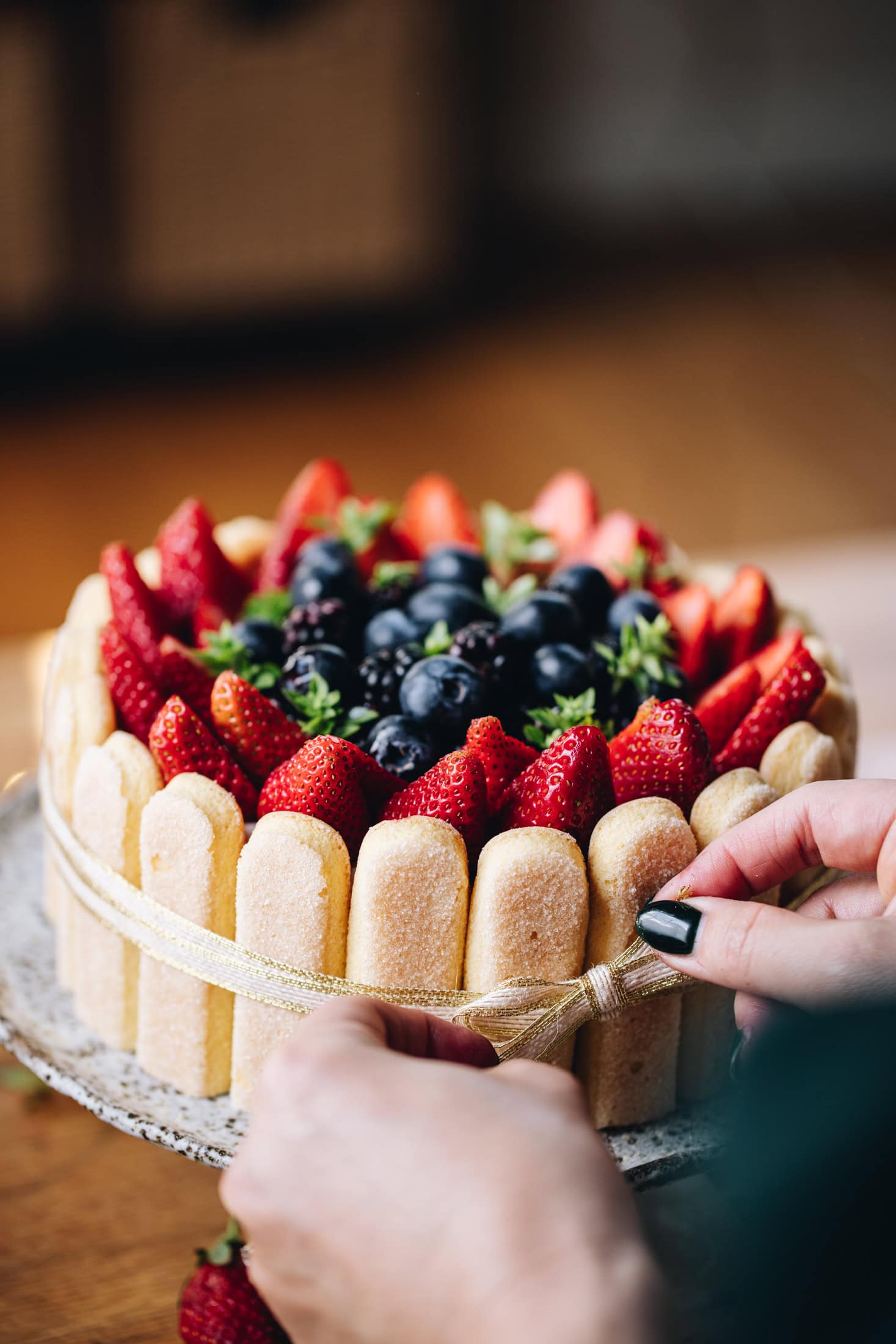 A no-bake Chocolate Charlotte Cake is decorated with berries. It is now being tied with a gold ribbon, hands are tying the bow at the front. It is on a ceramic cake stand, on a wooden bench. 