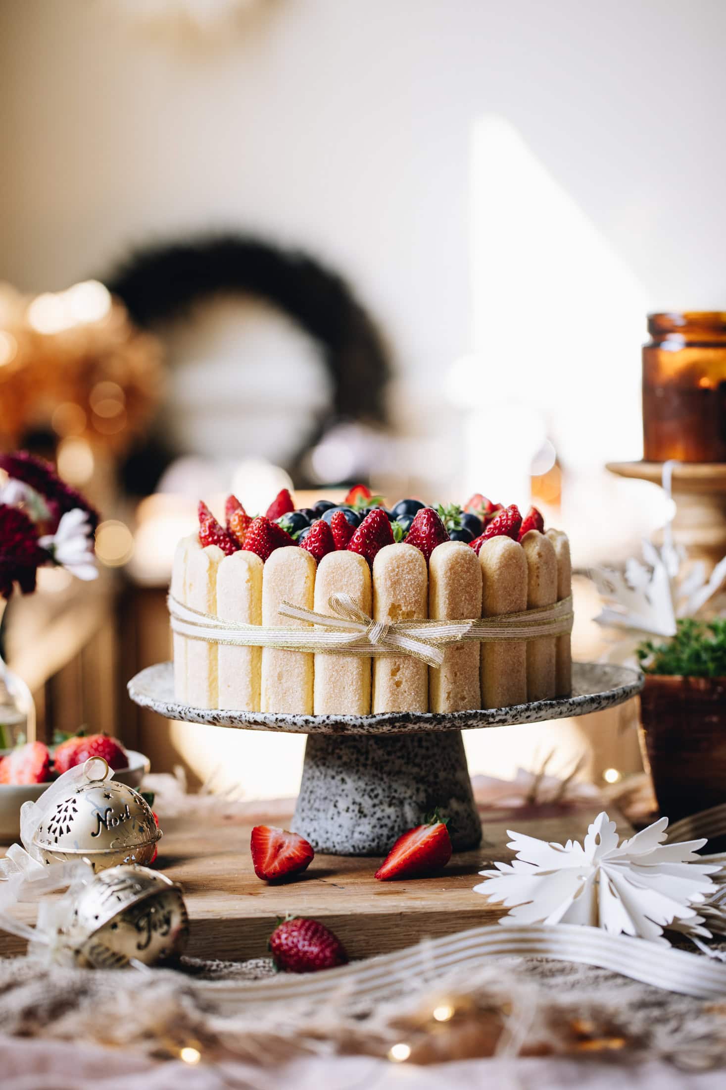 A no-bake Chocolate Charlotte Cake is decorated with berries. It is been tied with a gold ribbon, with a bow at the front. It is on a ceramic cake stand, on a wooden bench. It is surrounded by candles and Christmas decorations.