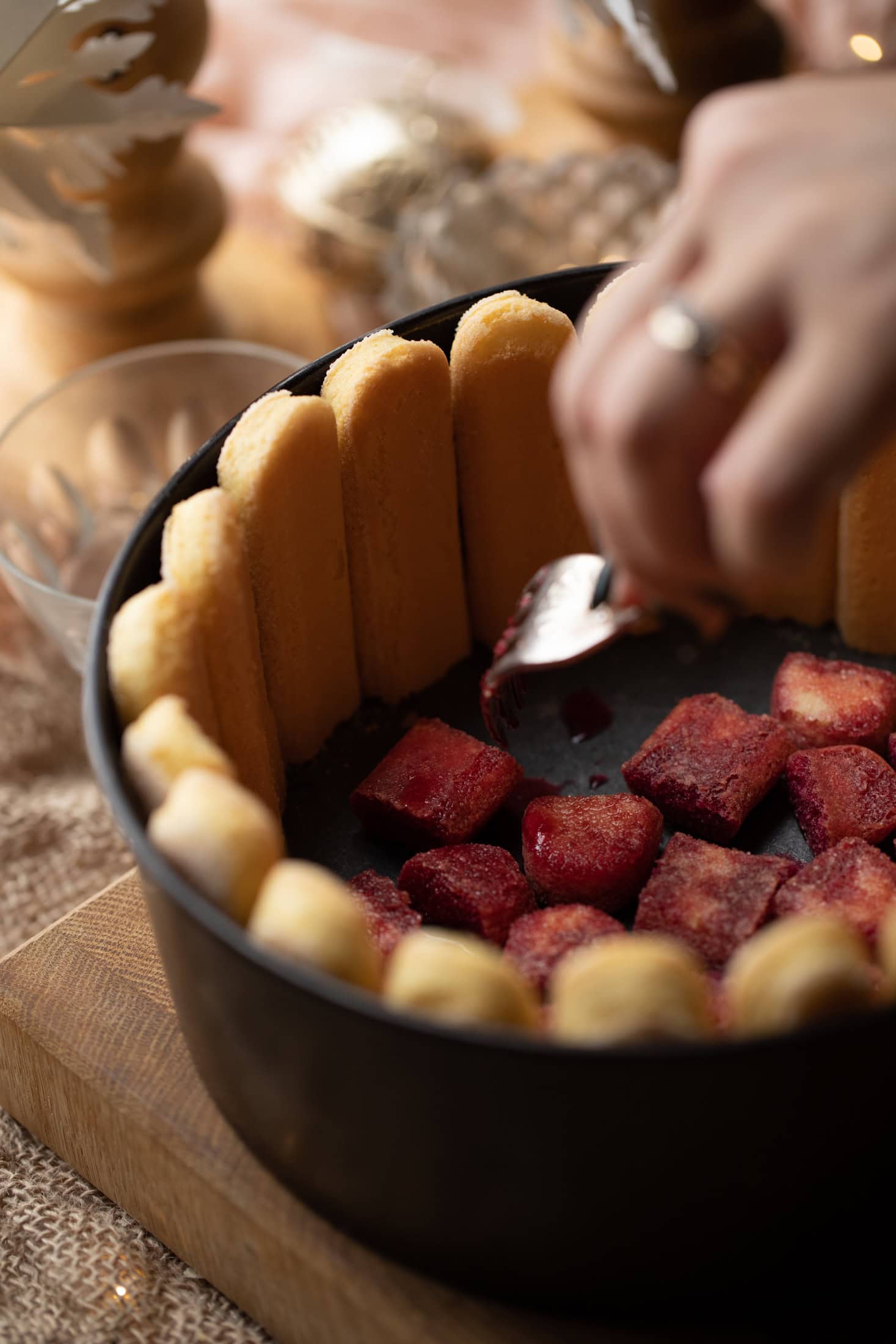 Lady fingers line a black cake tin with the bottoms cut off. In the base a fork is moving the lady Finger ends that are soaked in plum juice. It is sitting on a wooden board. 