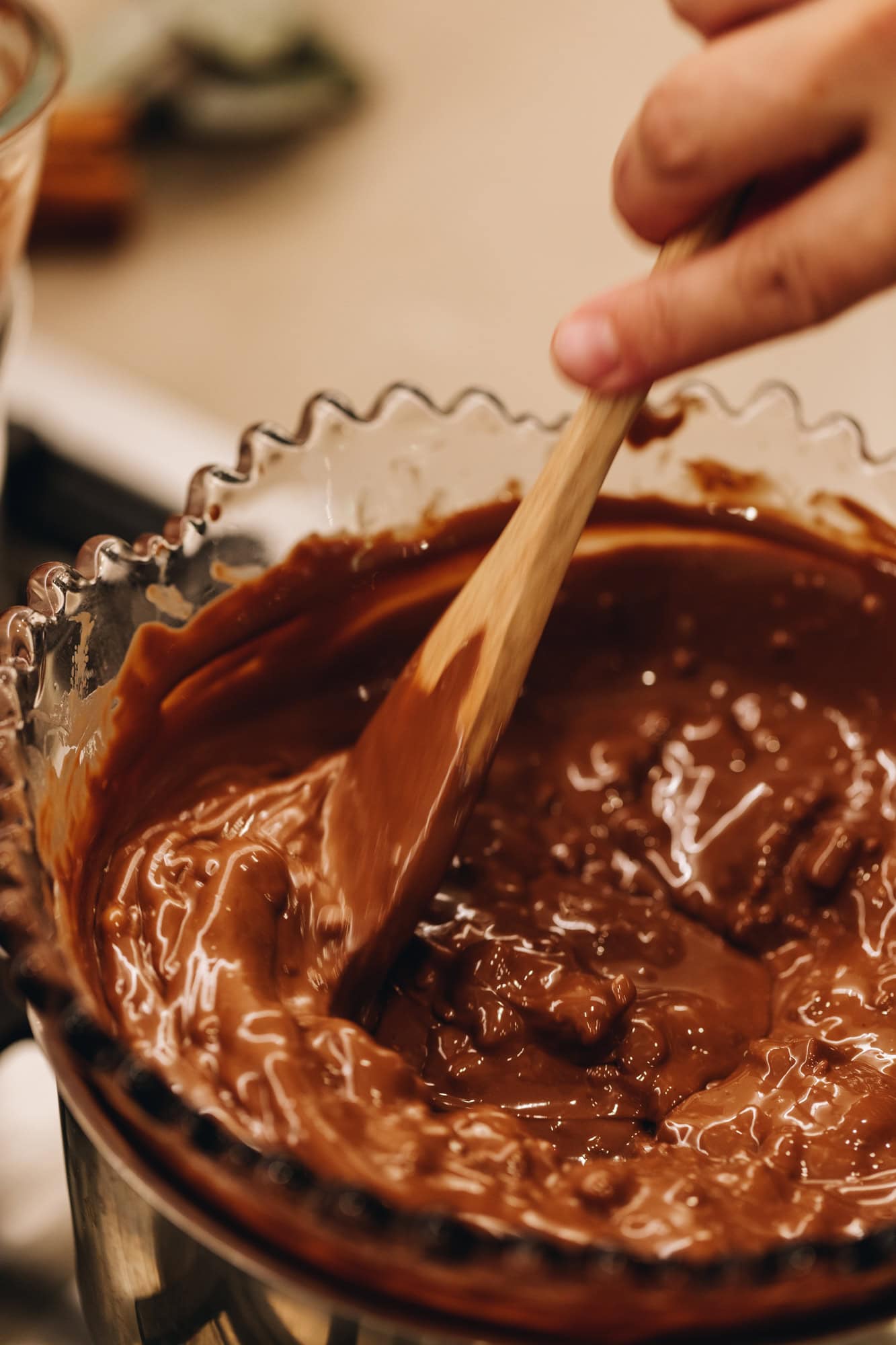 A vintage bowl of milk chocolate is melting over a pot of barely simmering water. It is being stirred by a wooden spoon. 