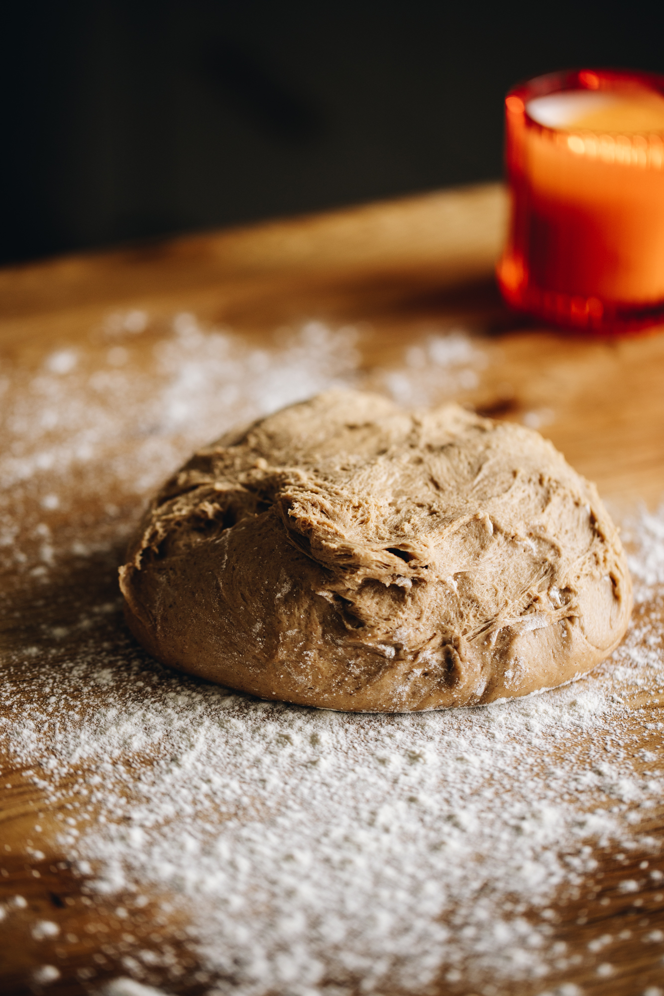 On a floured wooden table is a heavily spiced dough that has been kneaded and is in a ball. An orange candle is in the background. 