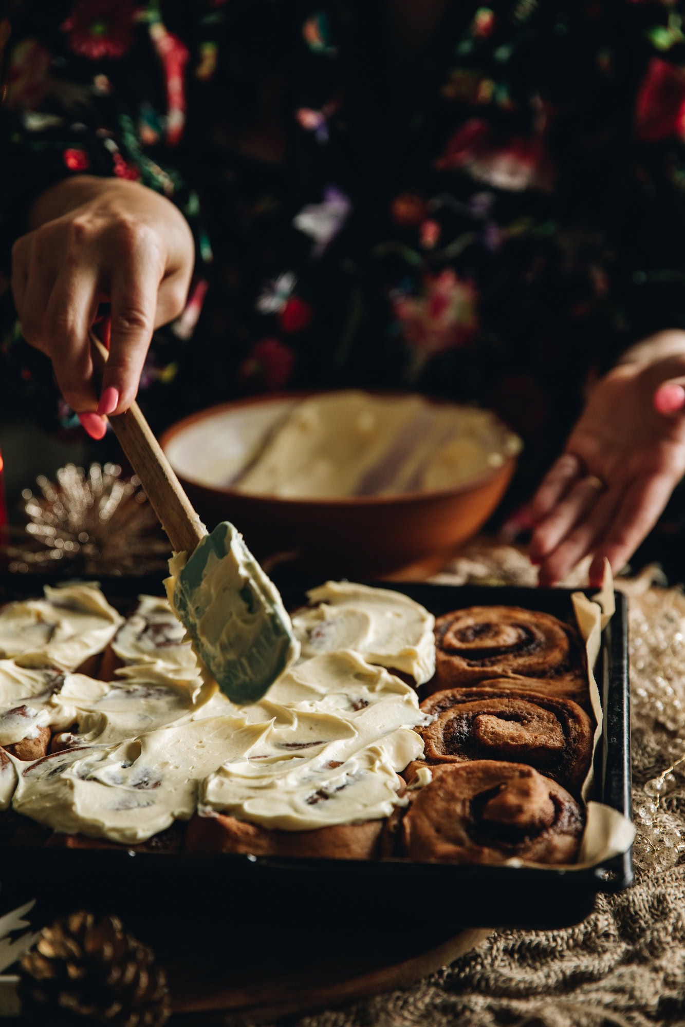 On a black tray are freshly baked sticky gingerbread scrolls. They are being smeared with cream cheese frosting with a spatula. 