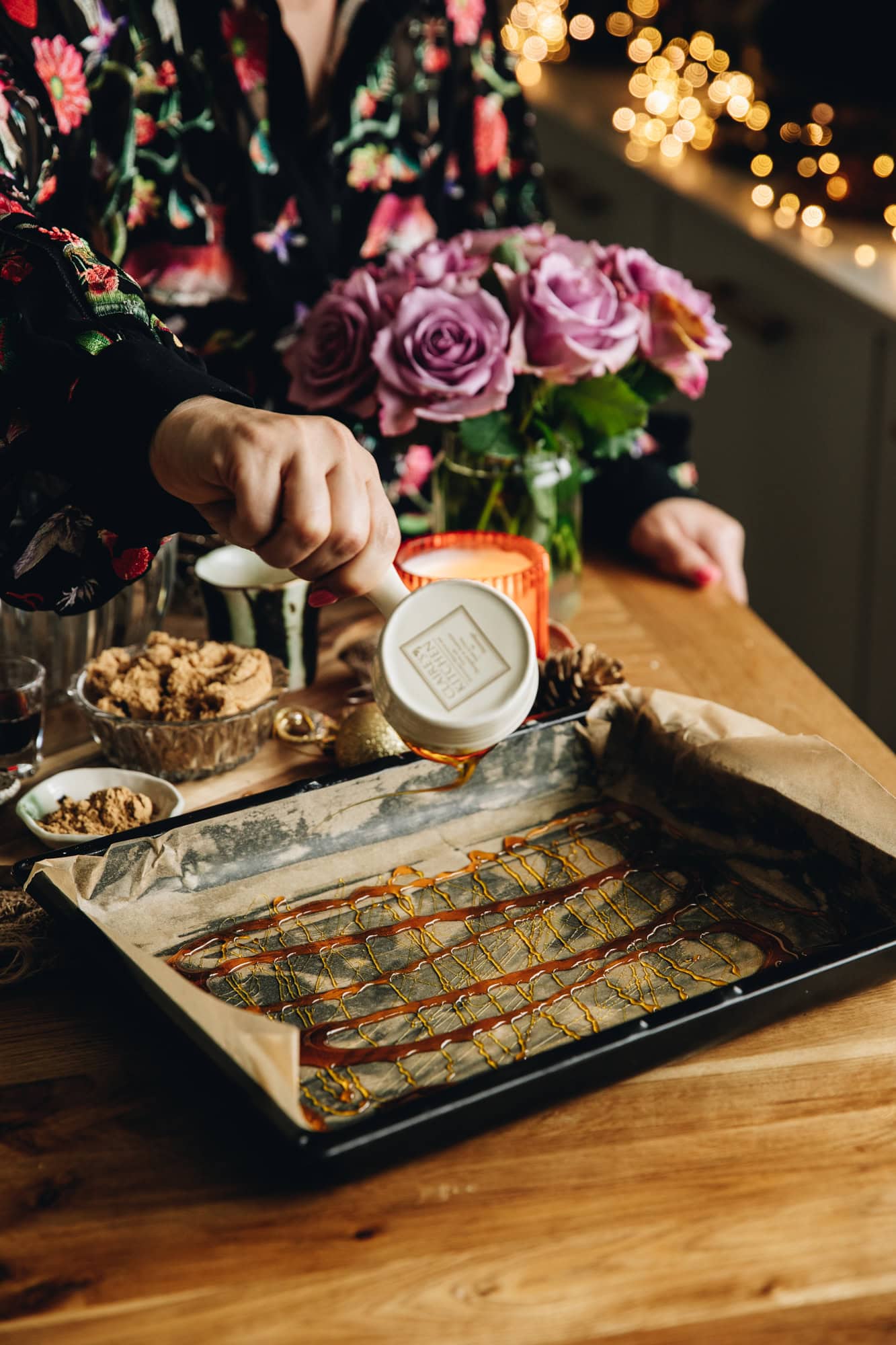 On a wooden table is a black tray lined with baking paper. It is being drizzled with golden syrup. Purple flowers sit in the background and fairy lights can be seen. 