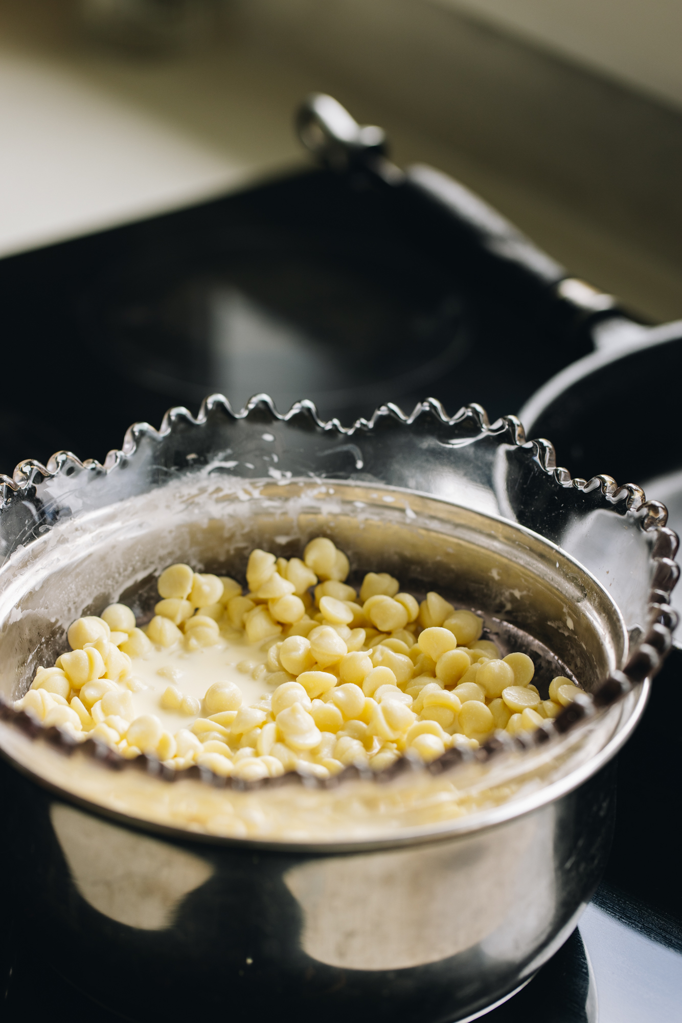 Chocolate drops are set over a double boiler in a vintage jagged edge bowl, sitting in a pot on a black stove.