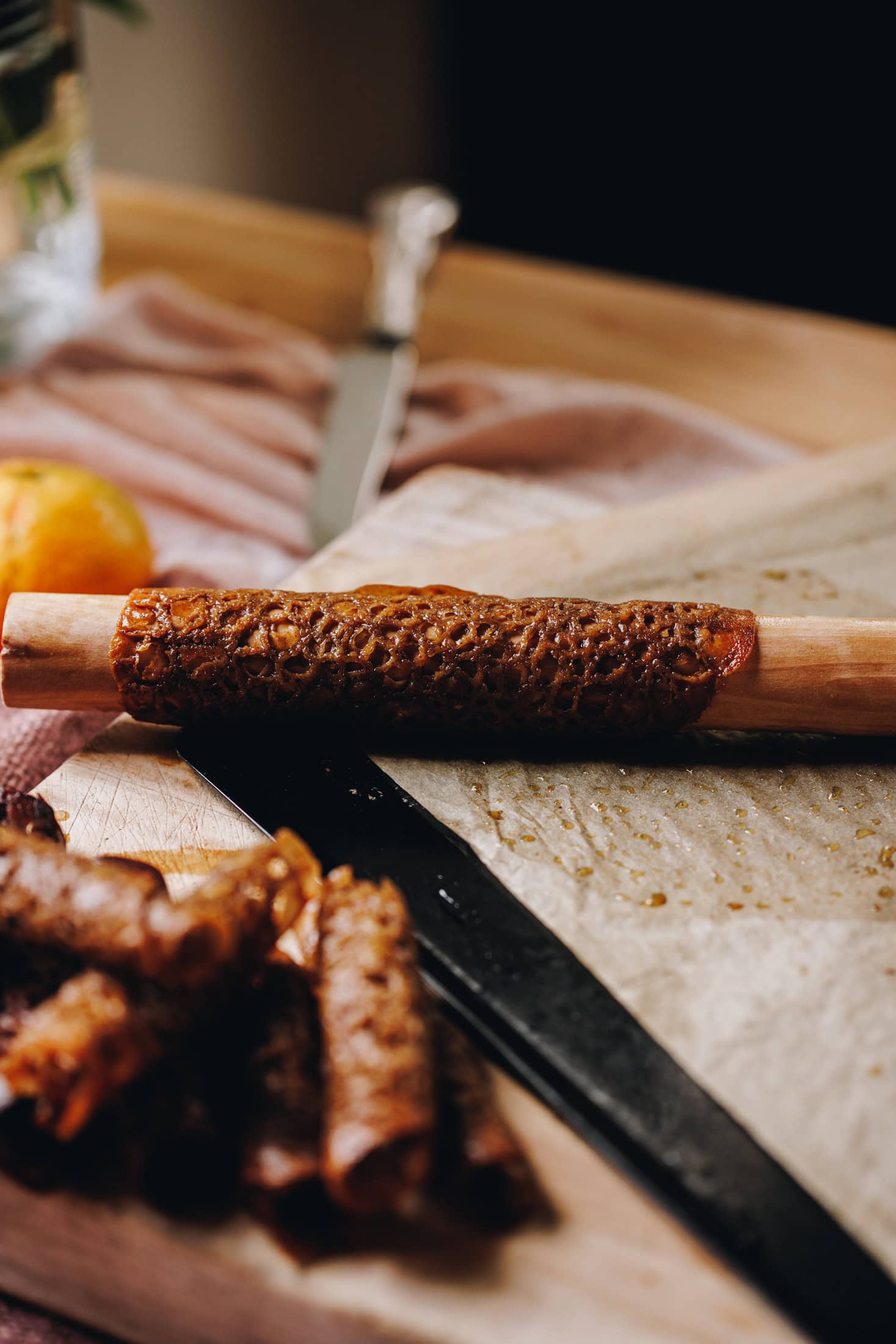 A brandy snapped is rolled upside a wooden handle. There is a pile of smaller Brandy snaps at the front of shot. It is on a wooden table.