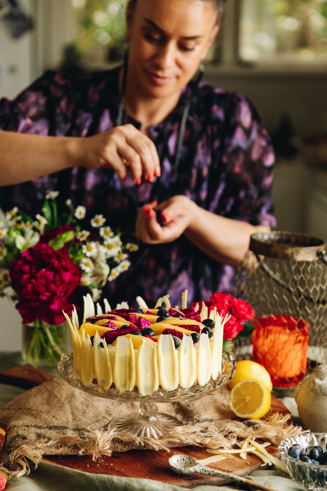 Naomi Toilalo standing behind a lemon and white chocolate cheesecake on a glass cake stand, styled with mango and dragon fruit and flowers.