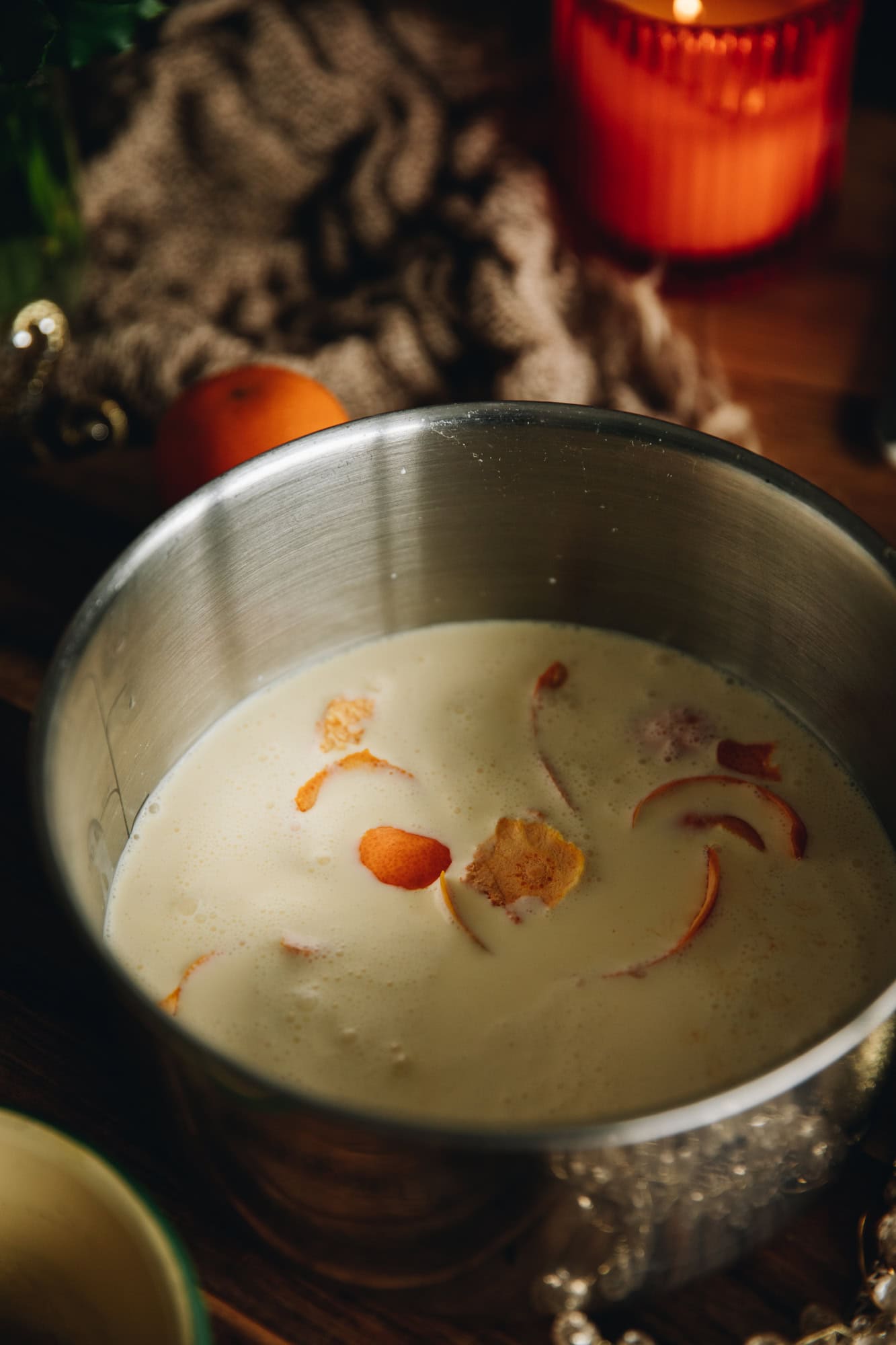 A silver pots sits on a wooden table and in the cream is orange peel. An orange candle burns in the background along with a mandarin and natural fabrics. 