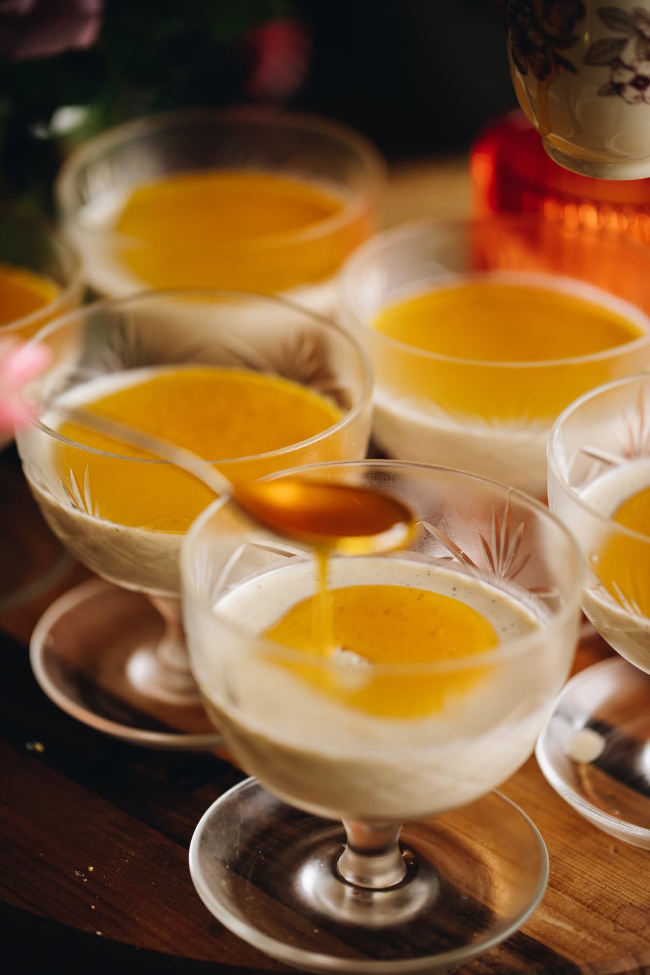 5 glass vintage ramekins sit on a wooden table. All of them have been filled with the cream mixture and four of them have orange syrup added on top. A teaspoon is drizzling the syrup on to the fifth panna cotta. An orange candle burns in the background.