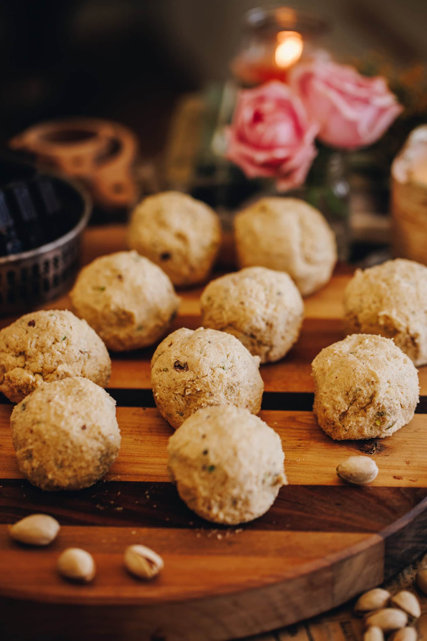 On striped wooden board sits 10 cookie dough rolled in to balls. There is a candle burning and pink flowers in the background.