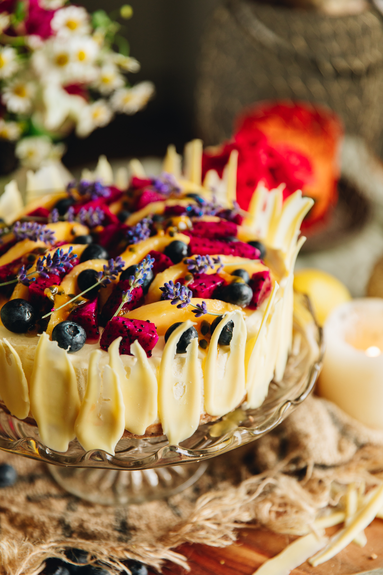 Lemon and white chocolate cheesecake with fruit and lavender topping and white chocolate decoration on a glass cake stand.