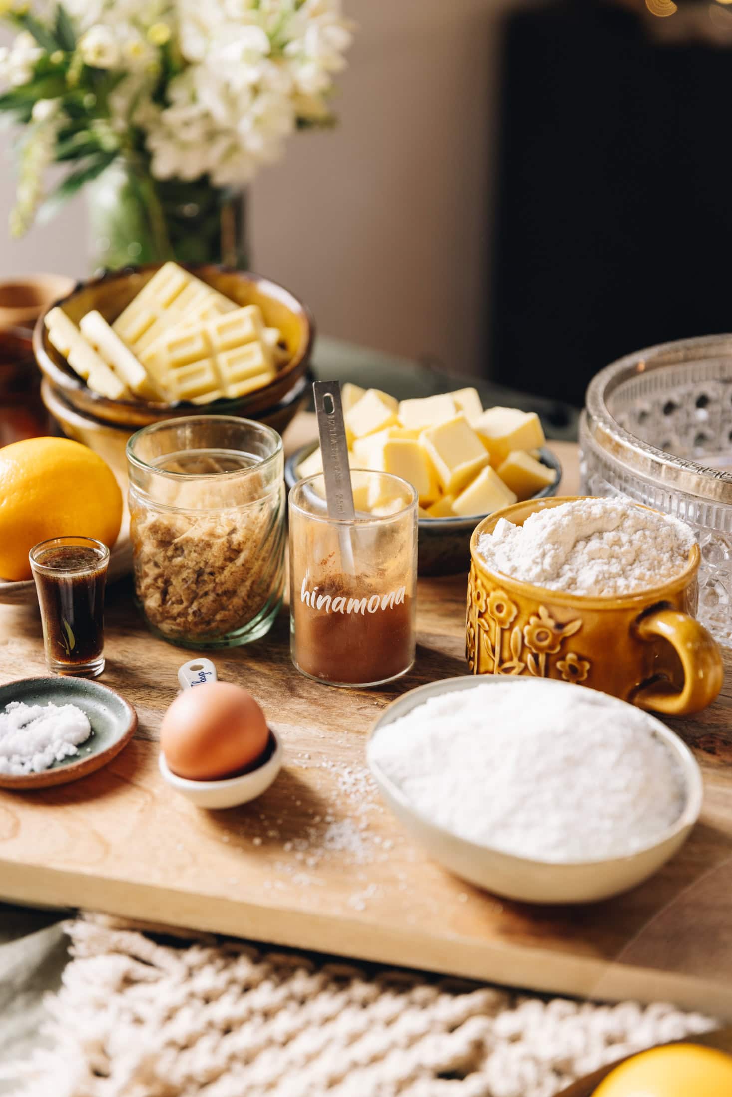 On a table sits a white weaved mat. On the mat is a wooden board that is filled with biscuit ingredients. There is sugar, hinamona, an egg, brown sugar, a lemon, white chocolate and flour in vintage cups and bowls. A vintage bowl and flowers are in the background. 