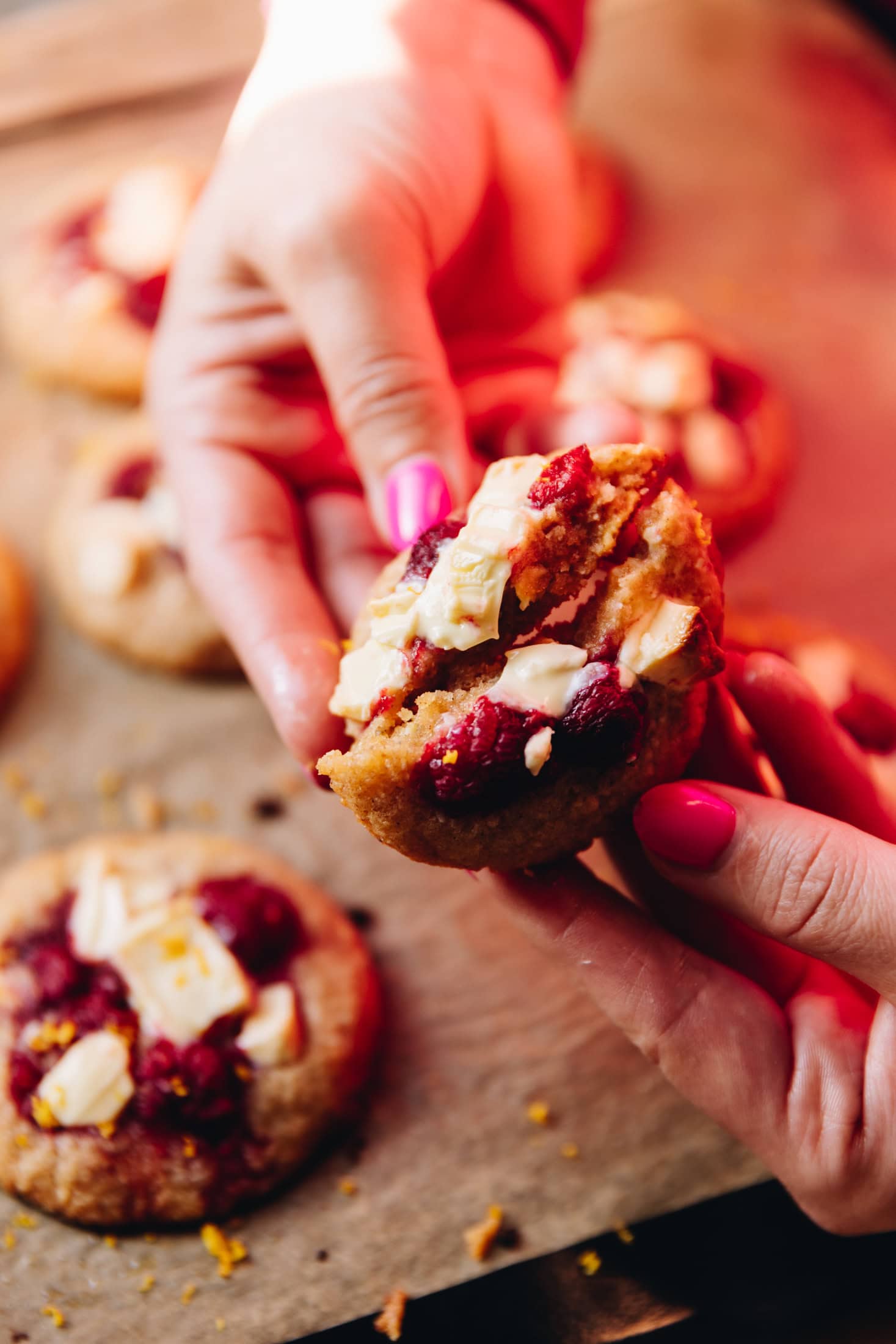 On a tray lined with brown baking paper is freshly baked white chocolate raspberry biscuits. Naomi is also holding one in her hands opening the cookie to reveal the inside.