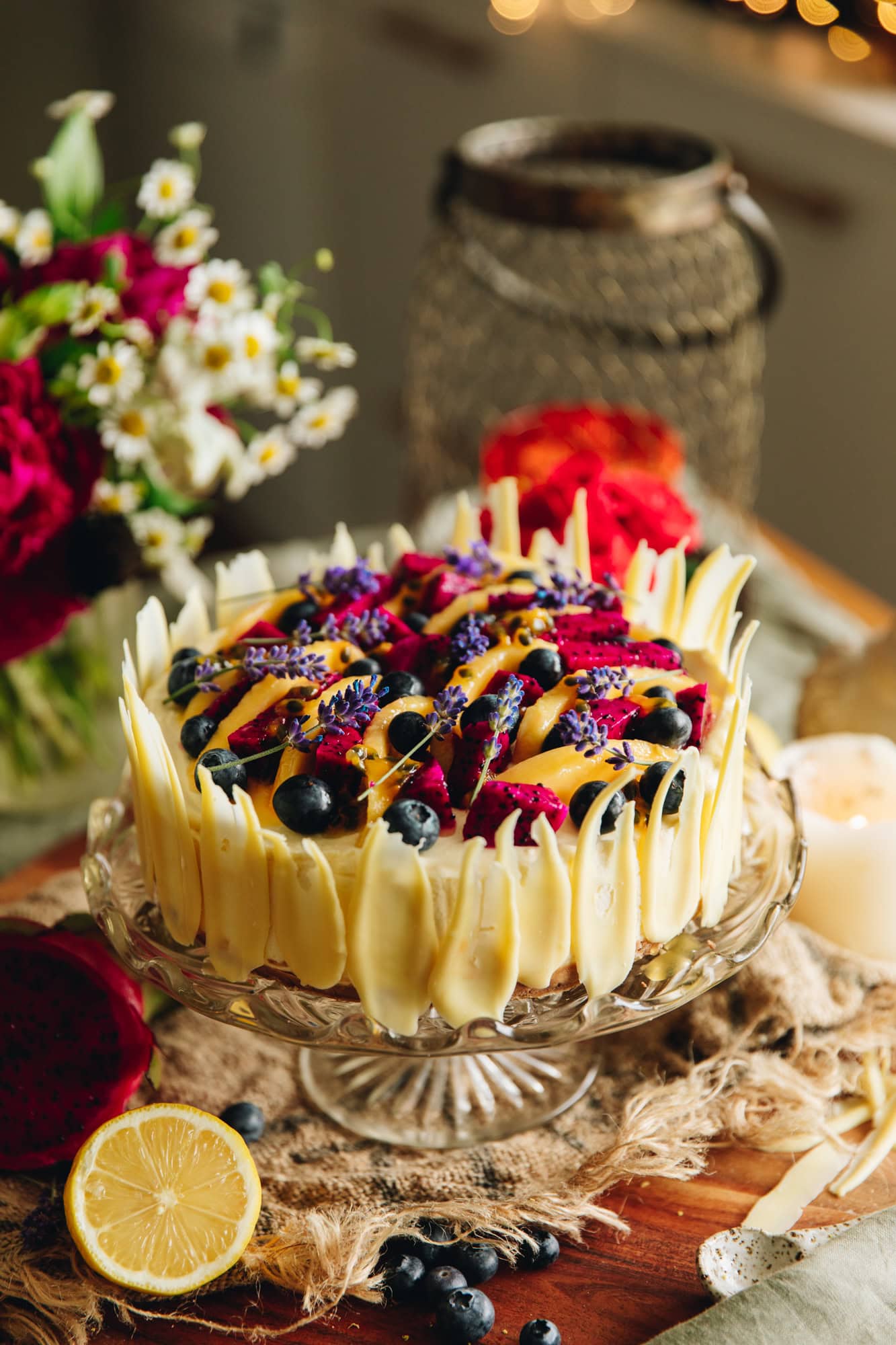 Lemon and white chocolate cheesecake on a glass cake stand with mango, dragon fruit, blueberries, and lavender flowers on top, styled on a rustic wooden board.