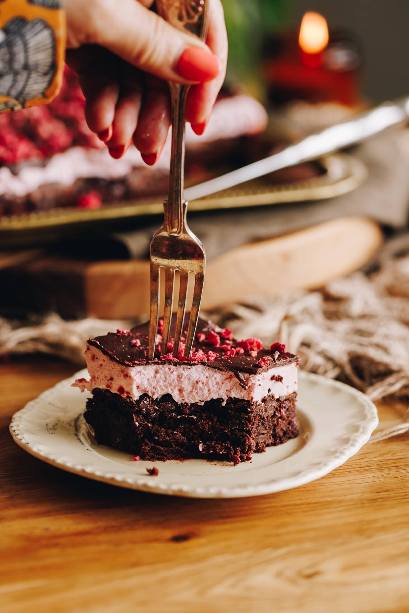 Slice of chocolate raspberry brownie with raspberry marshmallow topping on a vintage cream plate, being cut with a fork.