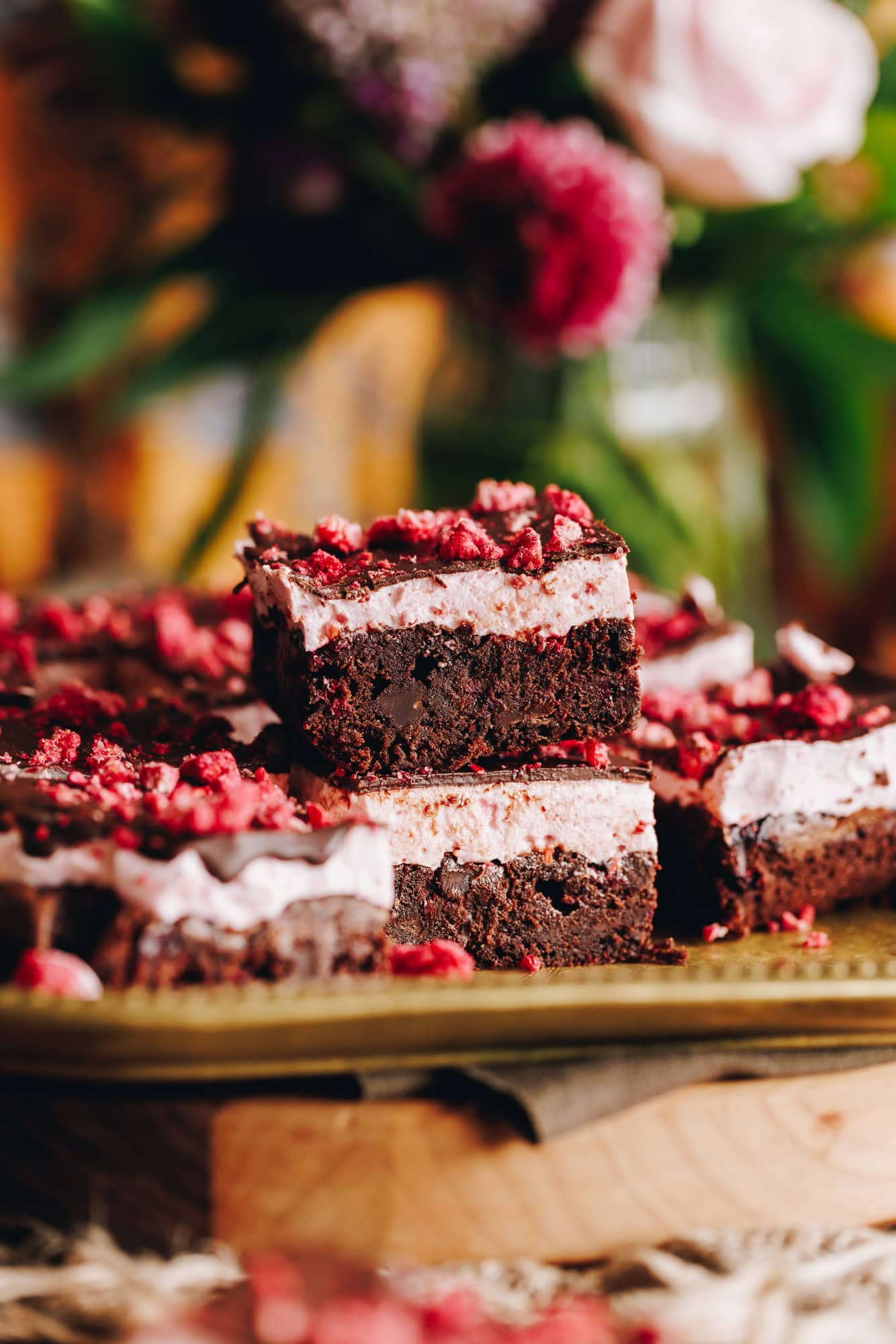 Slices of chocolate raspberry brownie with raspberry marshmallow arranged on a gold tray with flowers in the background.