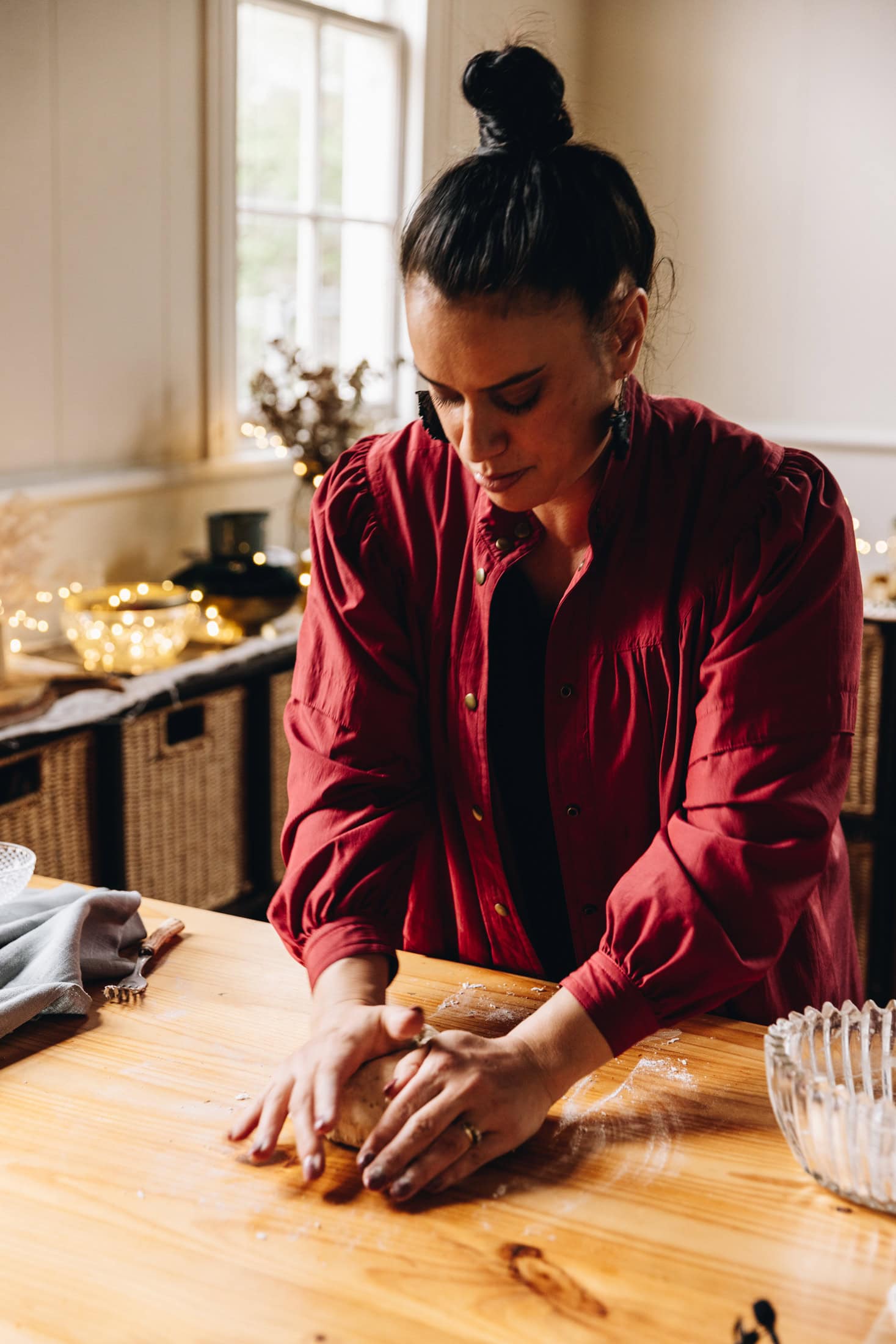 Naomi Toilalo is standing at a wooden table and is kneading dough for the Crunchy Yeasted Savoury Pastry. There is a tablecloth next to her and fairy lights in the background.