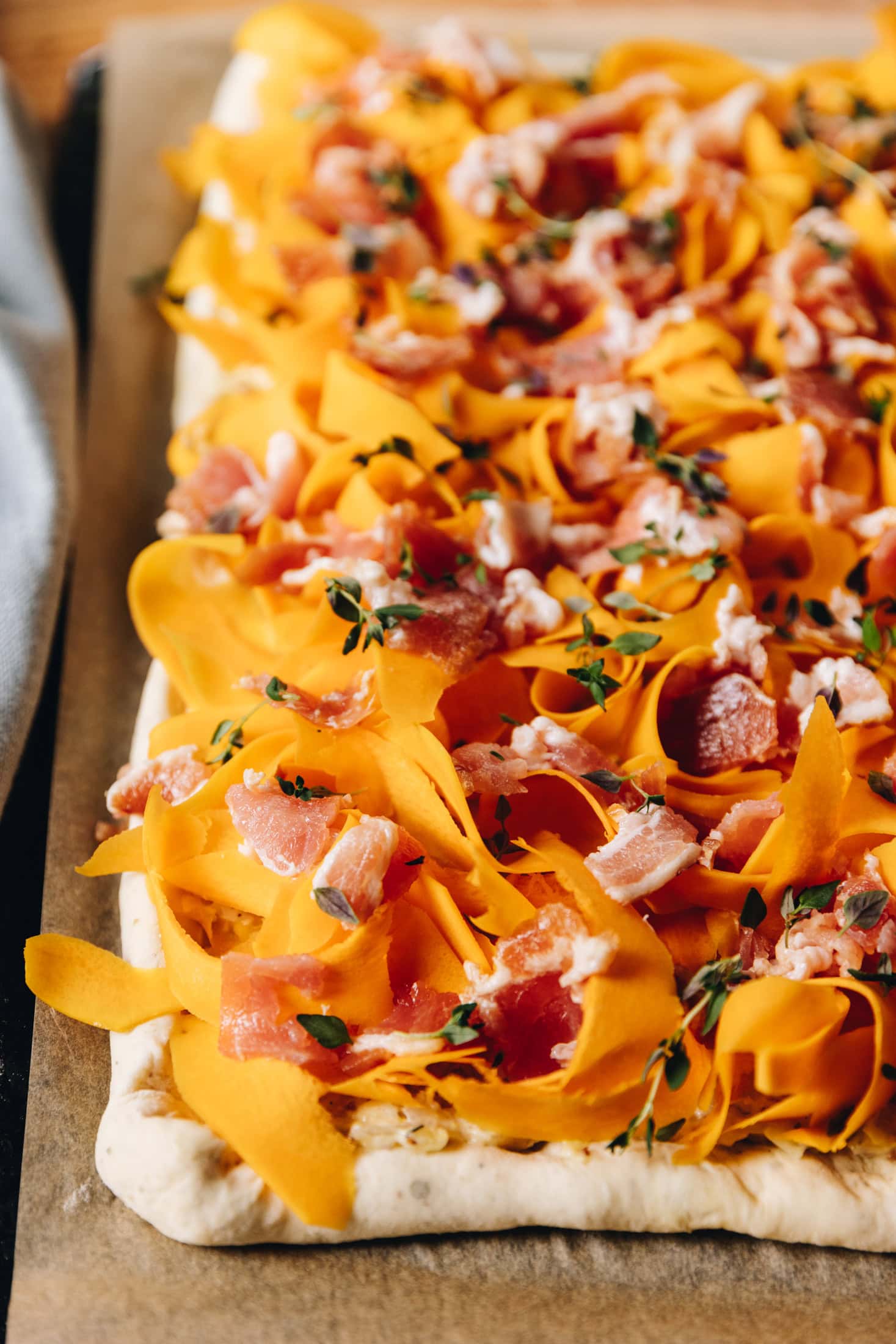 An unbaked Creamy Pumpkin tart is on a black tray lined with baking paper. It has shavings of pumpkin covering that whole tart with small strips of streaky bacon, fresh thyme leaves and a little bit of grated cheese on top. 