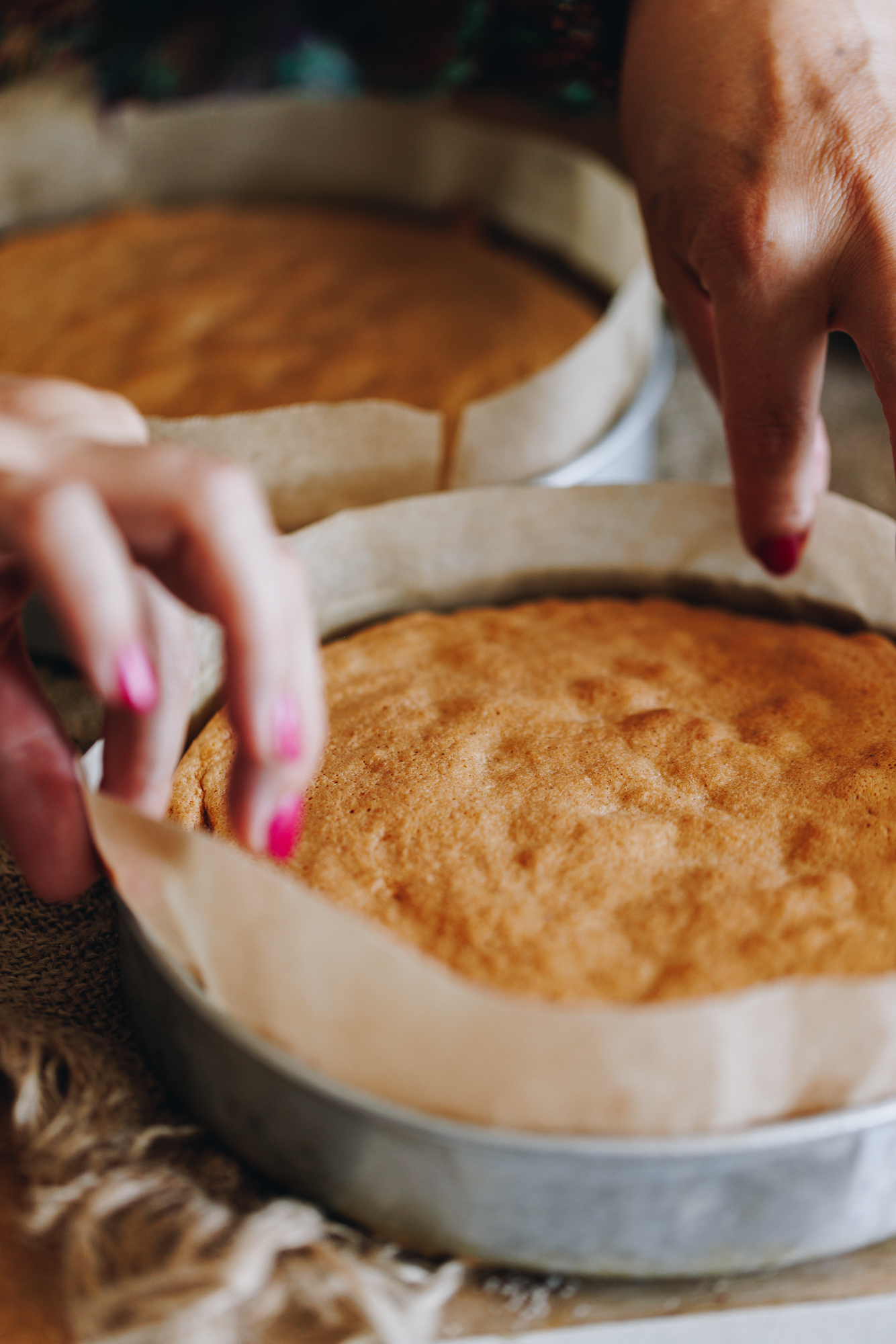 On a wooden bench sit two freshly baked gluten-free almond sponges, a hand is pulling the baking paper off the sides of one of the cakes. 