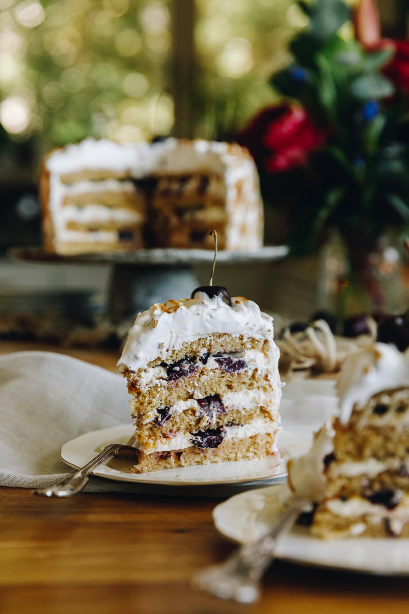 On a wooden table sits a slice of gluten0free almond sponge cake with layers of cream and cherries and four layers of sponge. A cherry sits on top. The whole cake cut open is blurred in the background. 