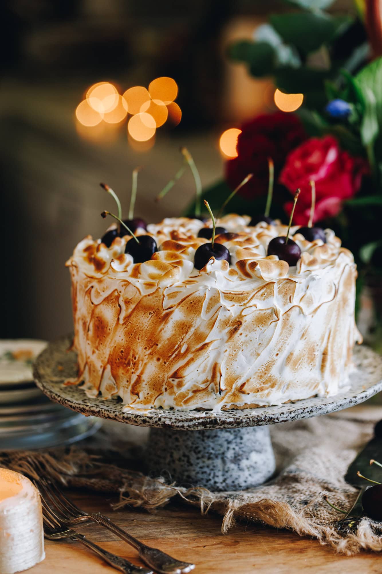 The decorated Gluten-free Almond sponge is covered with Swiss meringue that has been torched, it has fresh cherries on top. It is on a ceramic cake stand, sitting on a wooden table with flowers in the background.