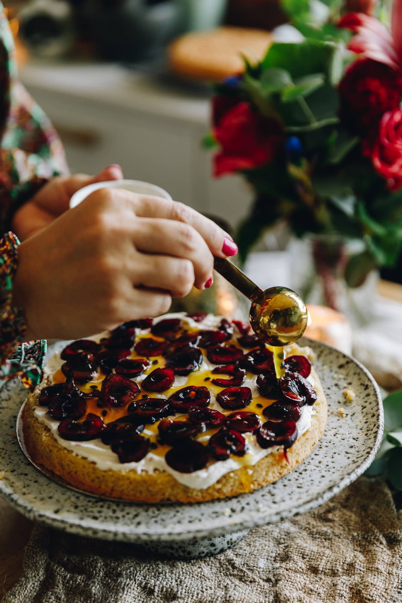 A ceramic cake stand is on top of a rustic piece of fabric. On the cake stand is one layer of the gluten-free almond sponge cake that has whipped cream and sliced cherries on it. A hand is drizzling passionfruit syrup over the cake.