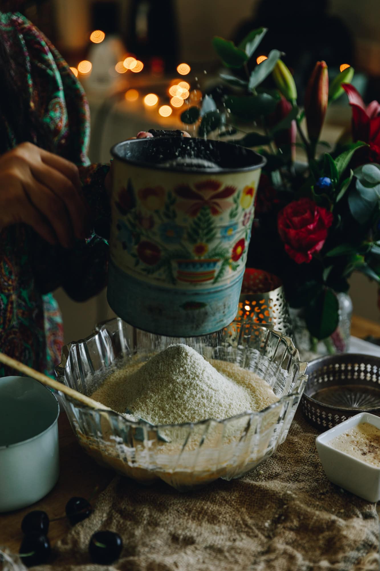 In a glass vintage bowl ground almonds are being sifted with a blue vintage sifter with a wooden handle. There is a candle burning in the background with flowers sitting on top a rustic piece of fabric.