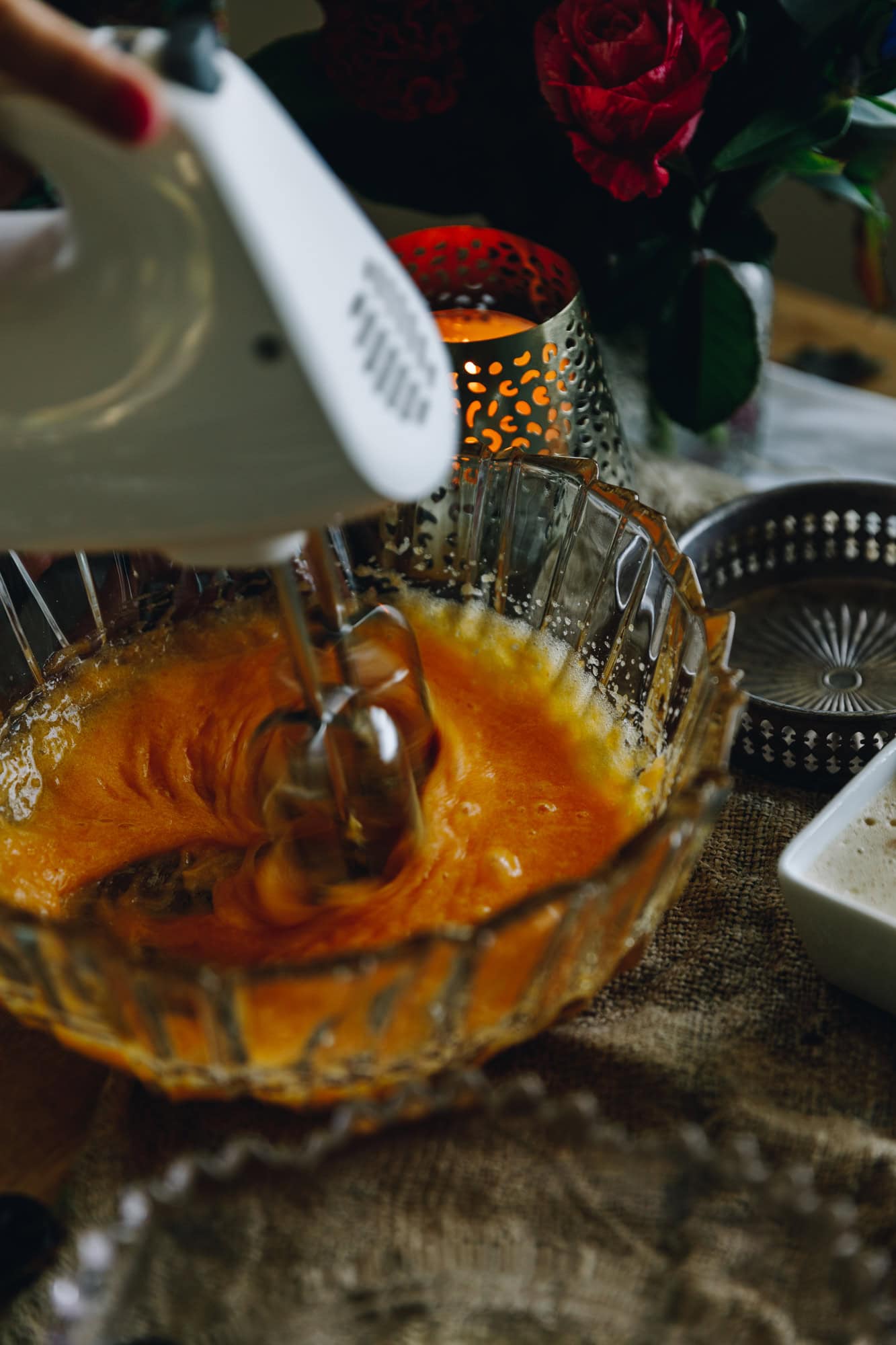 In a glass vintage bowl egg yolks are being whipped with a hand held beater. There is a candle burning in the background with flowers sitting on top a rustic piece of fabric. 
