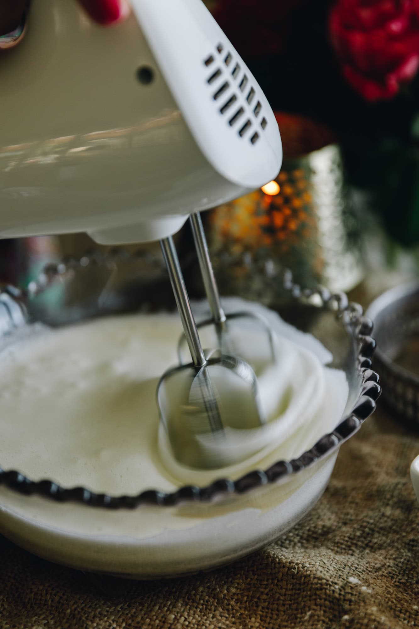 In a glass vintage bowl egg whites are being whipped with a hand held beater. There is a candle burning in the background with flowers sitting on top a rustic piece of fabric.
