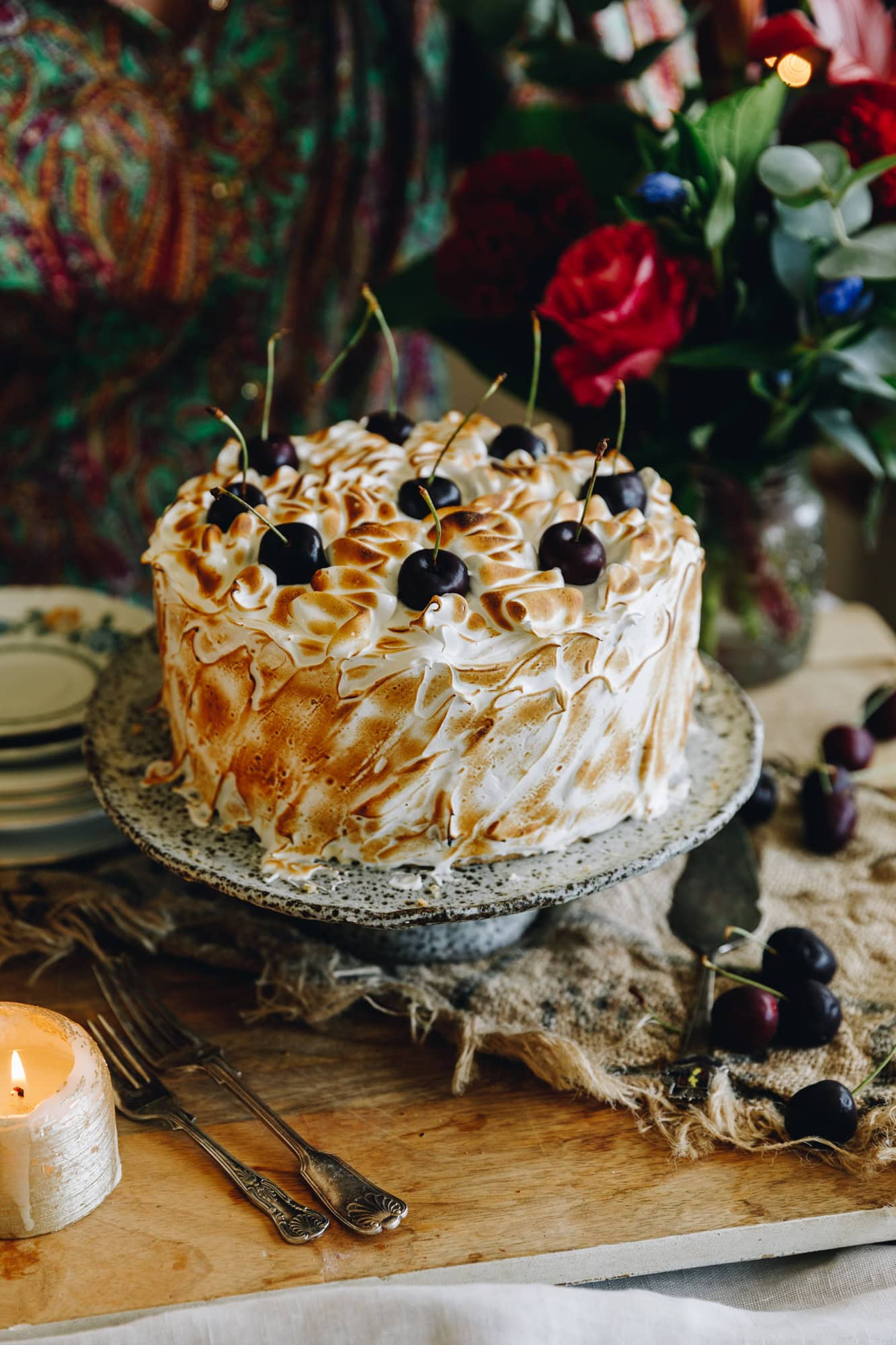 The decorated Gluten-free Almond sponge is covered with Swiss meringue that has been torched, it has fresh cherries on top. It is on a ceramic cake stand, sitting on a wooden table with flowers in the background.