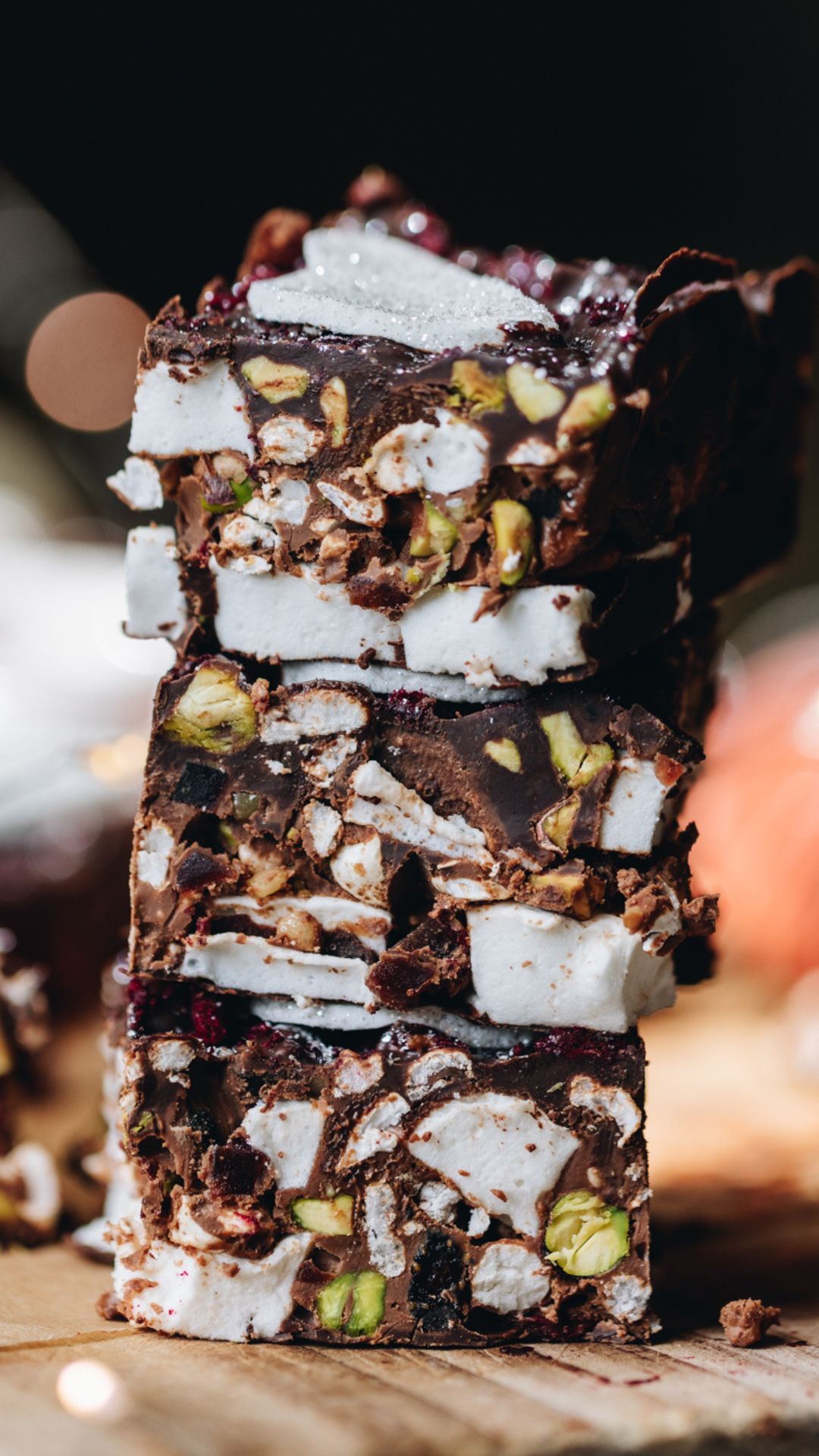 A wooden board has three pieces of homemade rocky road recipe on it. They have been stacked on top of each other and display the pistachio, vanilla marshmallow, strawberry pieces and chocolate rocky road. 