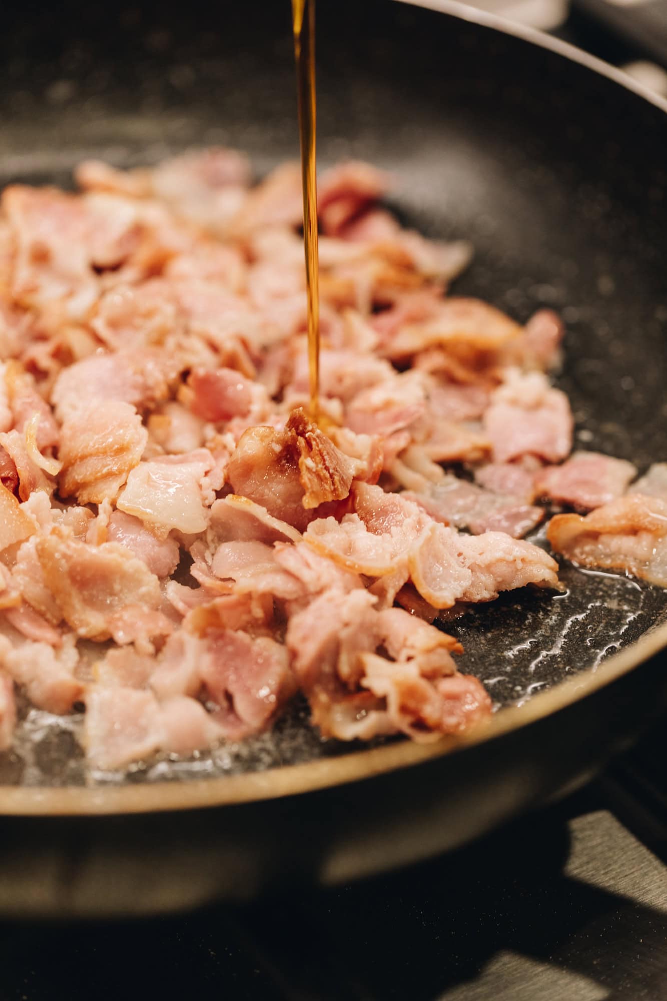 In a black frying pan is streaky bacon chopped I slices that has been cooked. Maple syrup is drizzling in to the pan at the same time. 