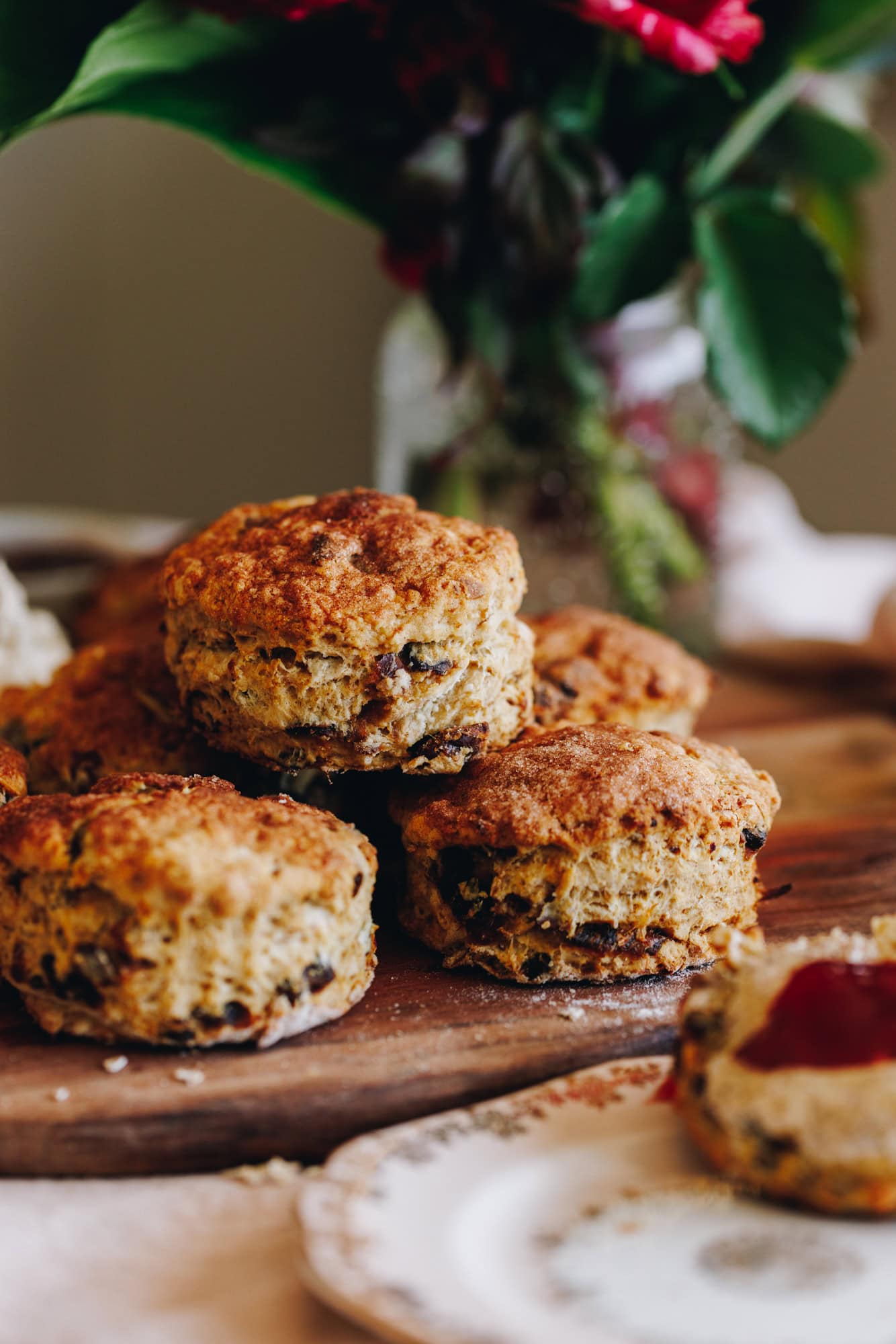 Freshly baked, round orange and date scones are piled on top of a wooden board. The dates are speckled through out the scone. In the corner is a pink vintage plate that has a scone with butter and jam on it. Flowers in na glass jar sit in the background. 