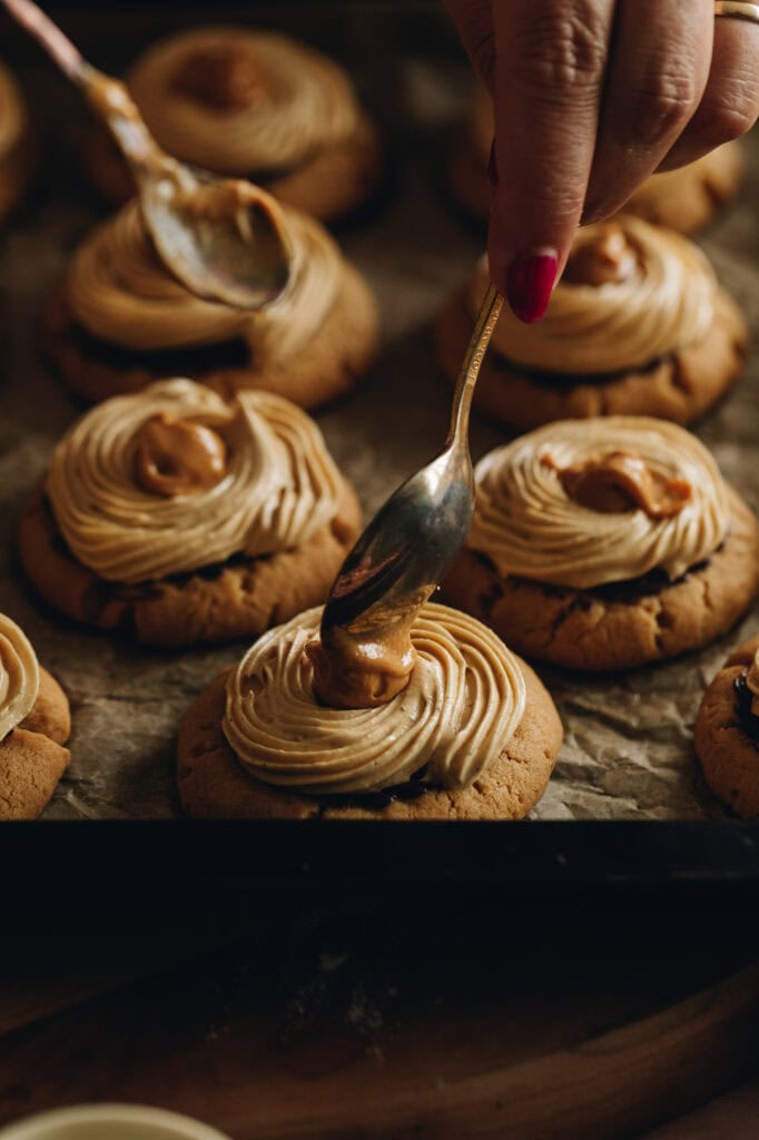 The baked Ultimate Peanut Butter Cookies are on a black tray with brown baking paper. The cookies have been piped with peanut butter buttercream and peanut butter is being placed in the centre with a teaspoon.