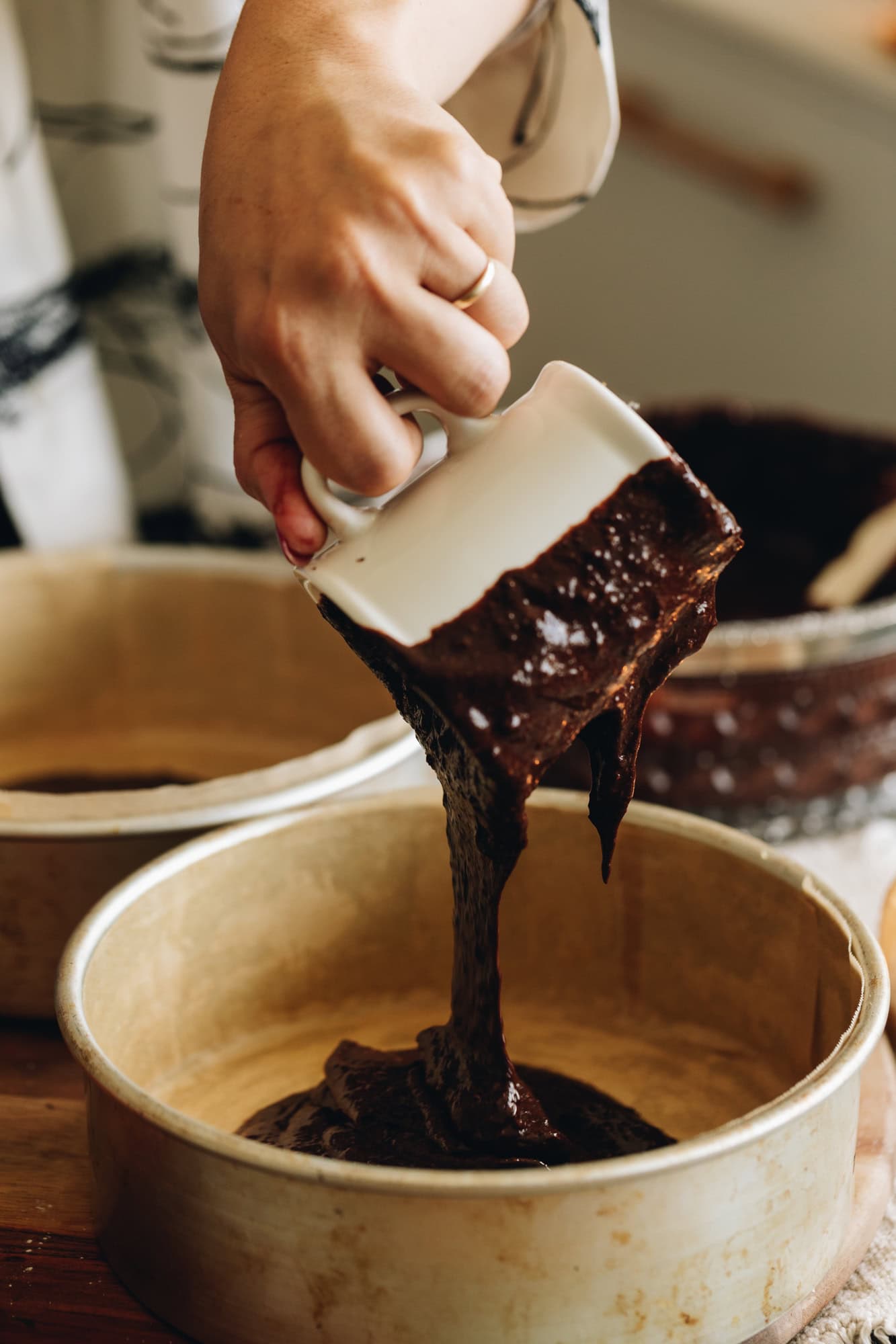 Two silver cake tins are sitting on a wooden board. They are lined with brown baking paper. In one tin a cup is pouring chocolate cake mixture in to it. 