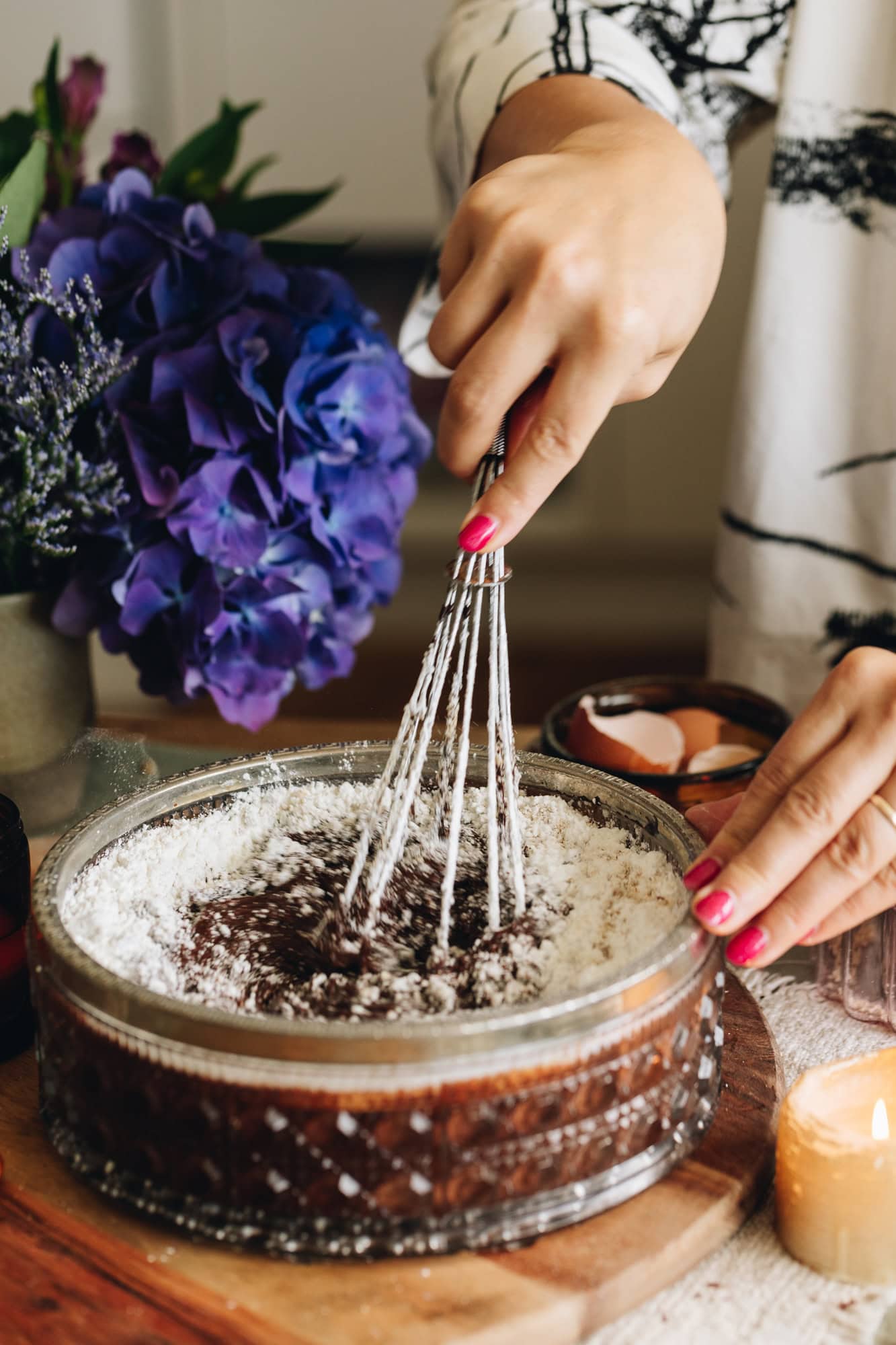 In a glass vintage bowl a chocolate batter and dry ingredients are being stirred with a wire whisk. It is sitting on a wooden bench with a purple flowers behind it. 