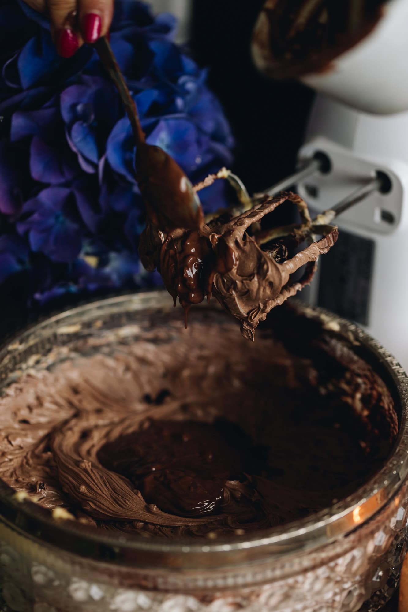 In a glass vintage bowl  is whipped chocolate butter mixture, in the centre melted chocolate has been added, it is also dripping off the edge of the hand beater whisk attachments. Purple flowers are in the background. 