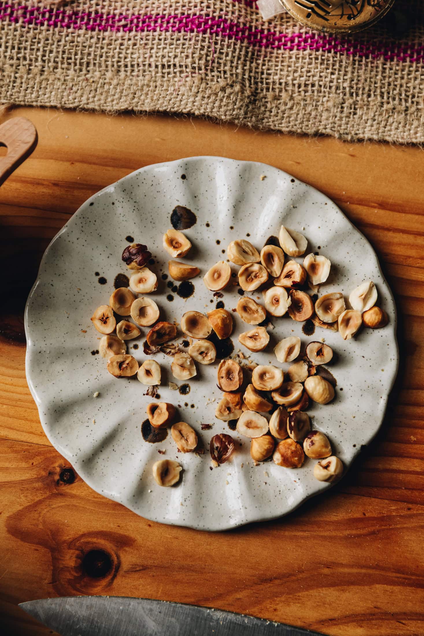 On a wooden table sits a ceramic plate with hazelnuts that have been toasted and cut in half. 