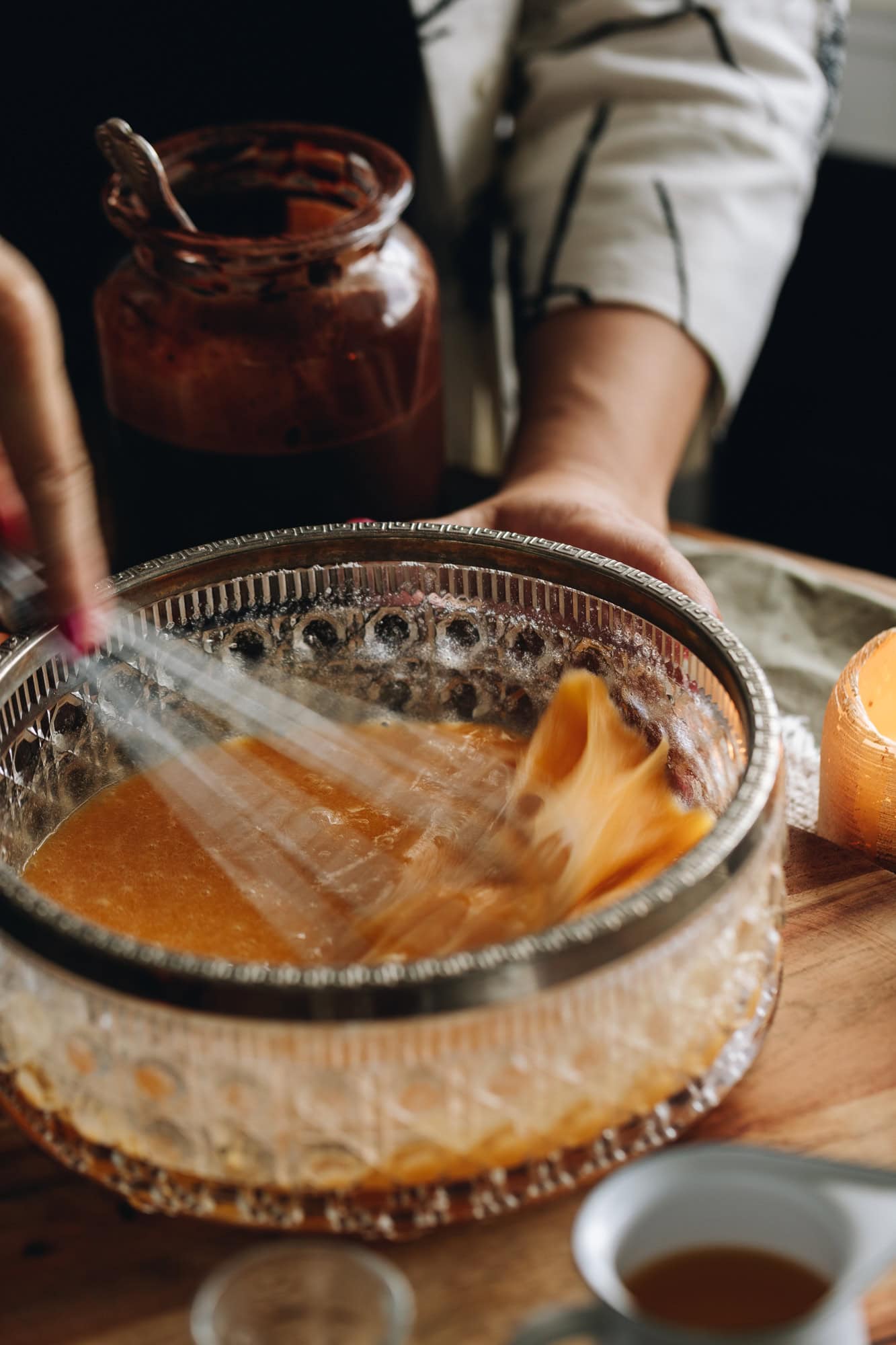 In a glass vintage bowl eggs are being whipped with a wire whisk. It is sitting on a wooden bench with a jar of cocoa mixture behind it. 