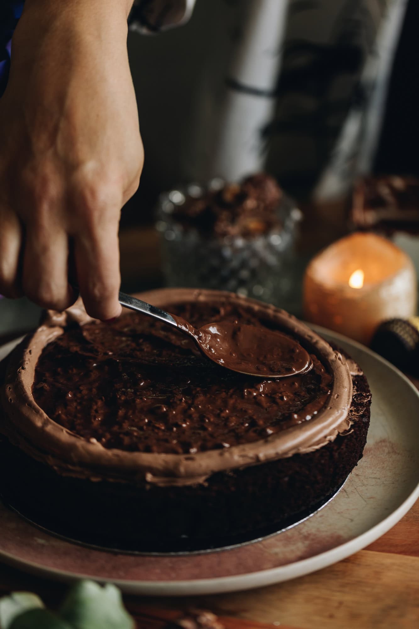 A pink plate is sitting on a wooden board. On the plate is a chocolate hazelnut cake that has the buttercream spread on it and a a border is piped around the edge of the cake. A spoon is spreading chocolate hazelnut spread in to the centre of the cake. A candle is burning in the background.