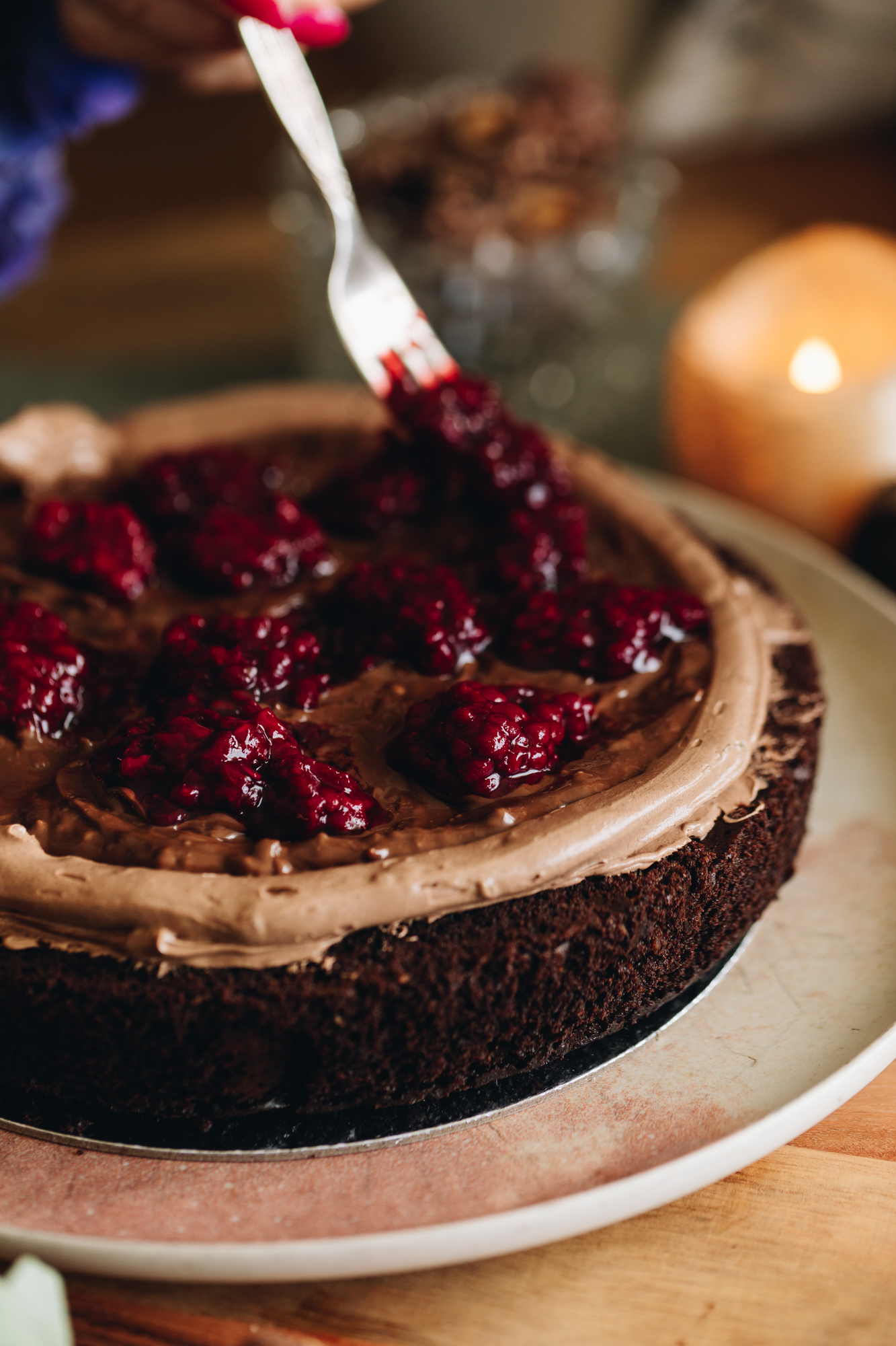 A chocolate hazelnut cake sits on a pink plate, on top of a wooden table. It is been topped with chocolate buttercream, chocolate hazelnut spread and is having canned boysenberries placed on it with a fork. 