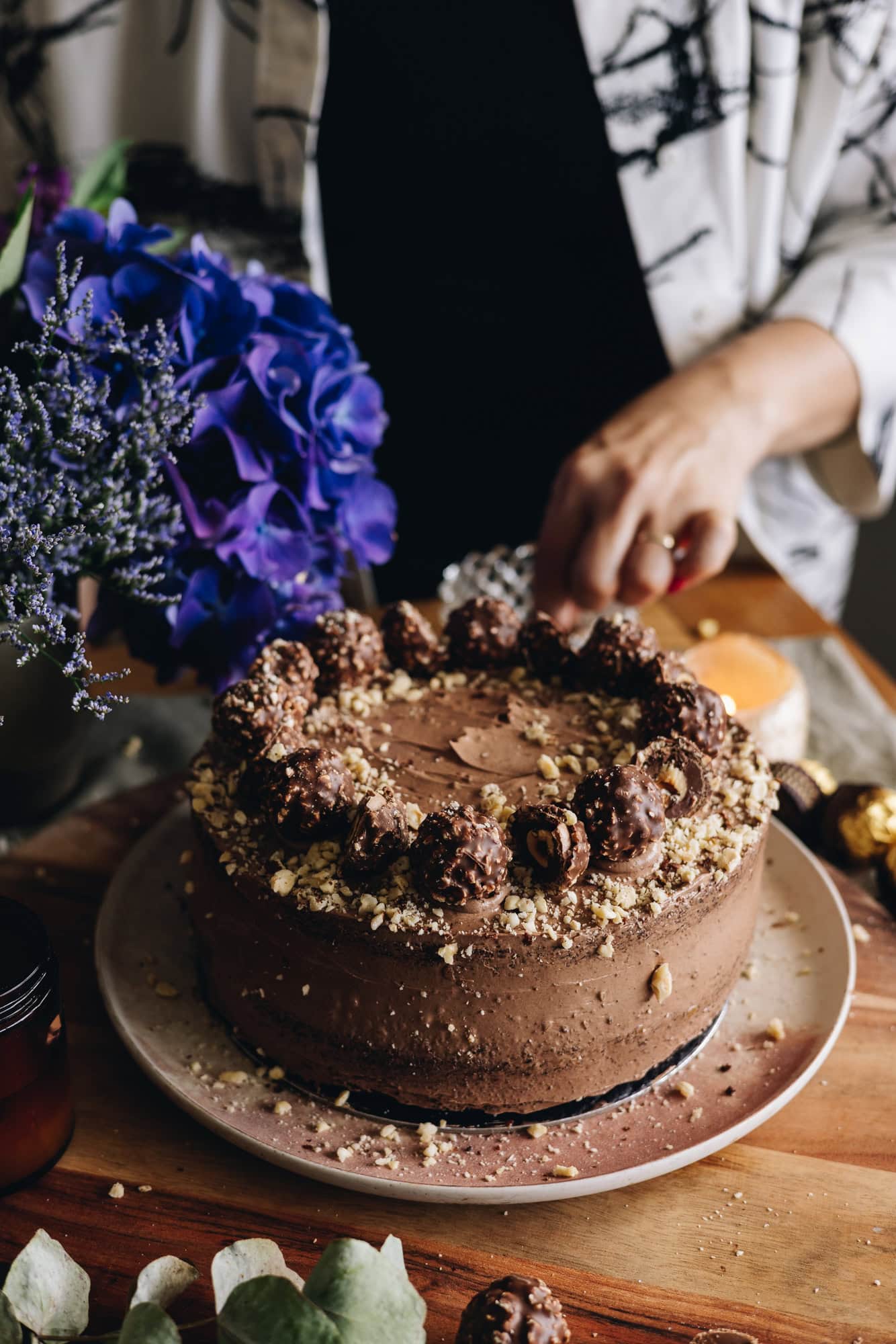 A whole chocolate hazelnut cake is sitting on a pink plate on a wooden table. It is covered in chocolate frosting with chopped hazelnuts and forerro rocher chocolates on top. It has purple flowers in the background. Hans are adding a decoration on to the cake.