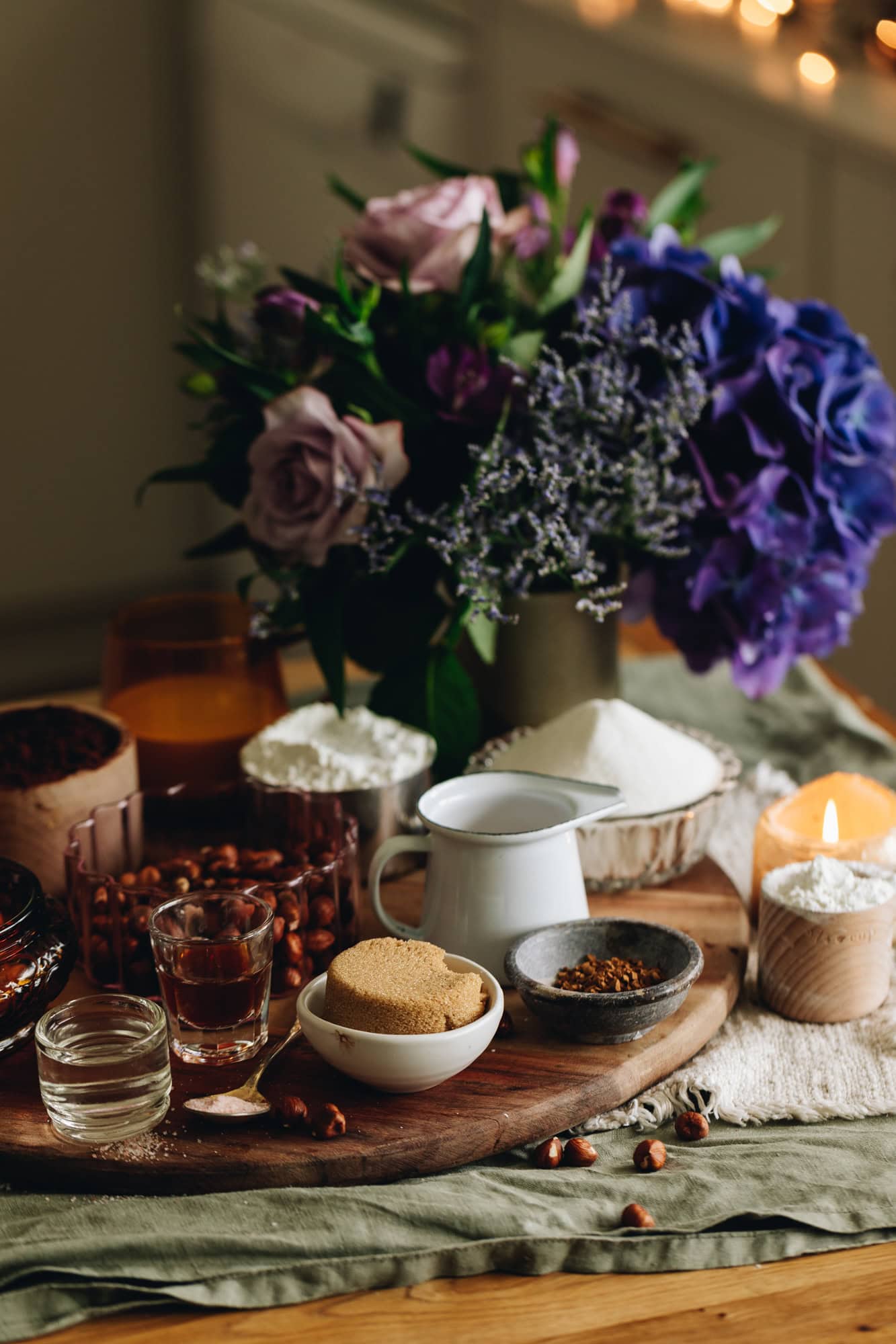Ingredients are in different vintage bowls and are sitting on a wooden board on top of wooden table There is a bunch of flowers next to the ingredients with a burning candle. 