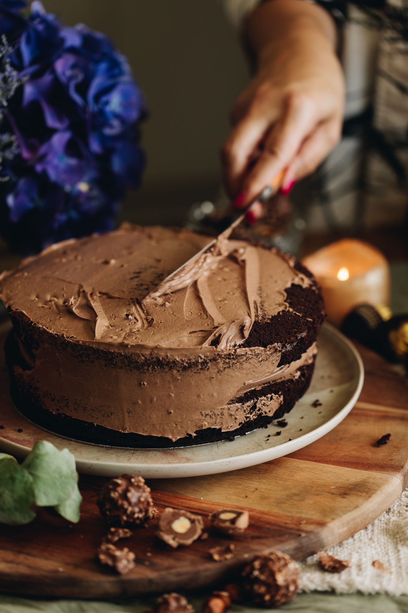 Two chocolate hazelnut cakes sit on a pink plate, on top of a wooden table. It is been topped with chocolate buttercream and is being smoothed out with a palette knife. 