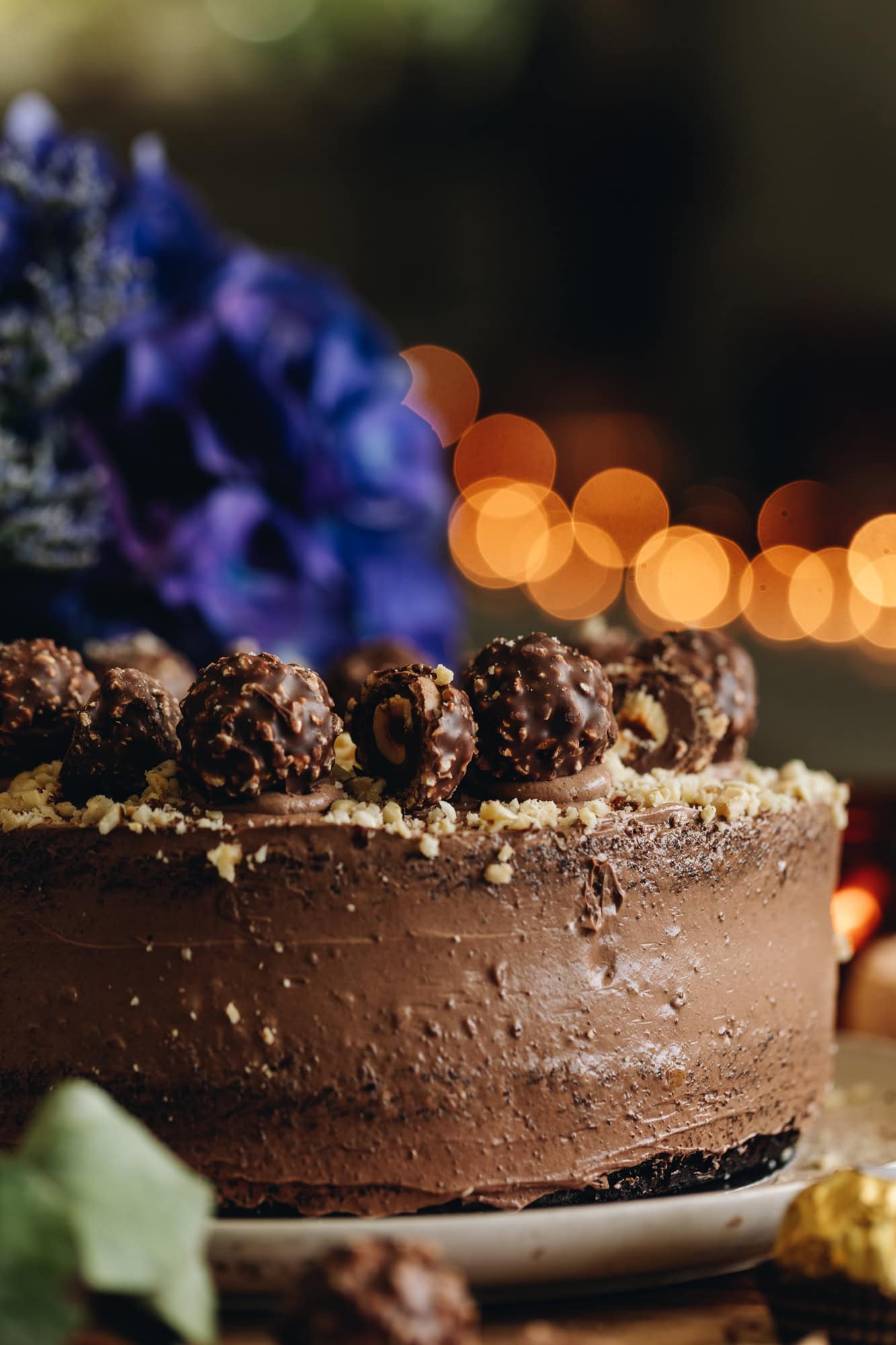 A close shot shows a whole chocolate hazelnut cake is sitting on a pink plate on a wooden table. It is covered in chocolate frosting with chopped hazelnuts and forerro rocher chocolates on top. It has purple flowers in the background.