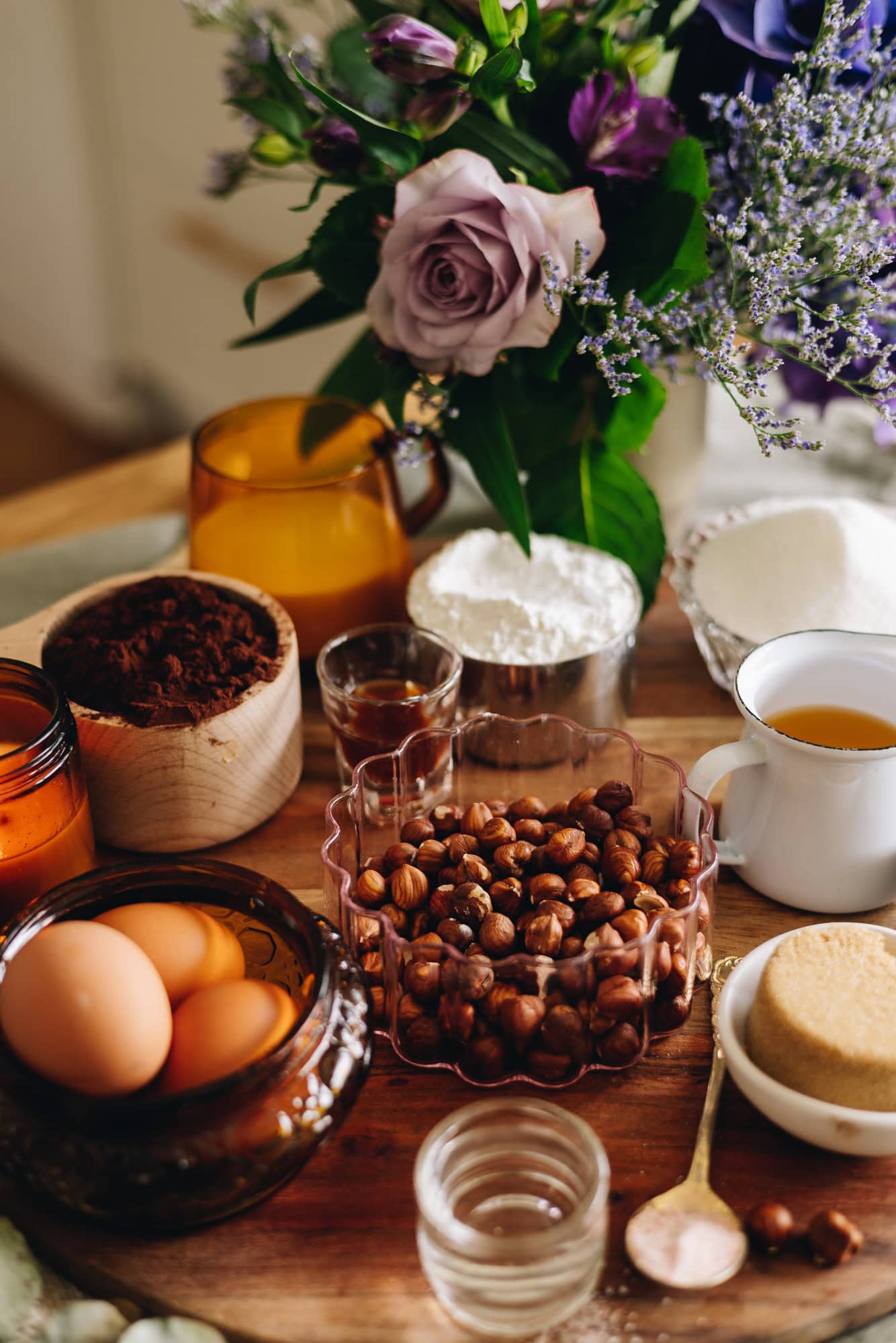 Cake ingredients are in different vintage bowls and are sitting on a wooden board on top of wooden table. There is a bunch of flowers next to the ingredients.