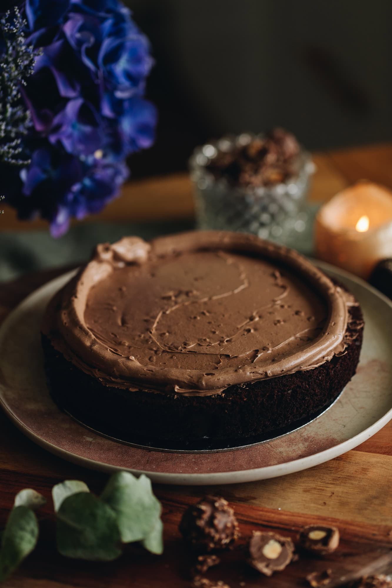 A pink plate is sitting on a wooden board with purple flowers behind it. On the plate is a chocolate hazelnut cake that has the buttercream spread on it and a a border is piped around the edge of the cake.